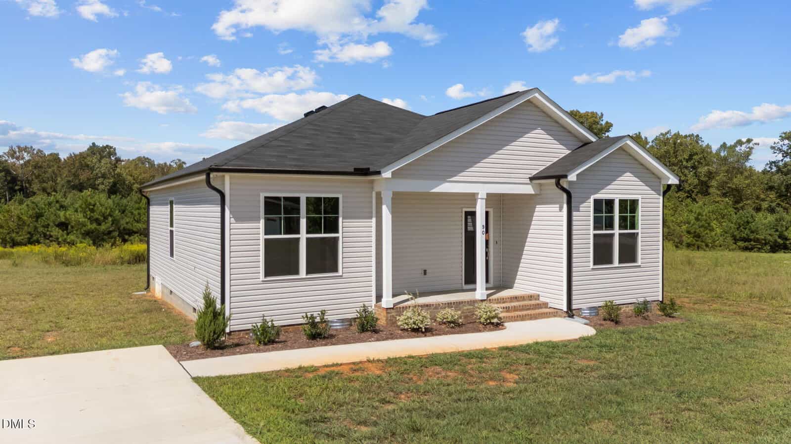 A small, modern single-story white house with a black roof and front porch, set on a grassy lot with young shrubs and trees in the background under a blue sky with scattered clouds.