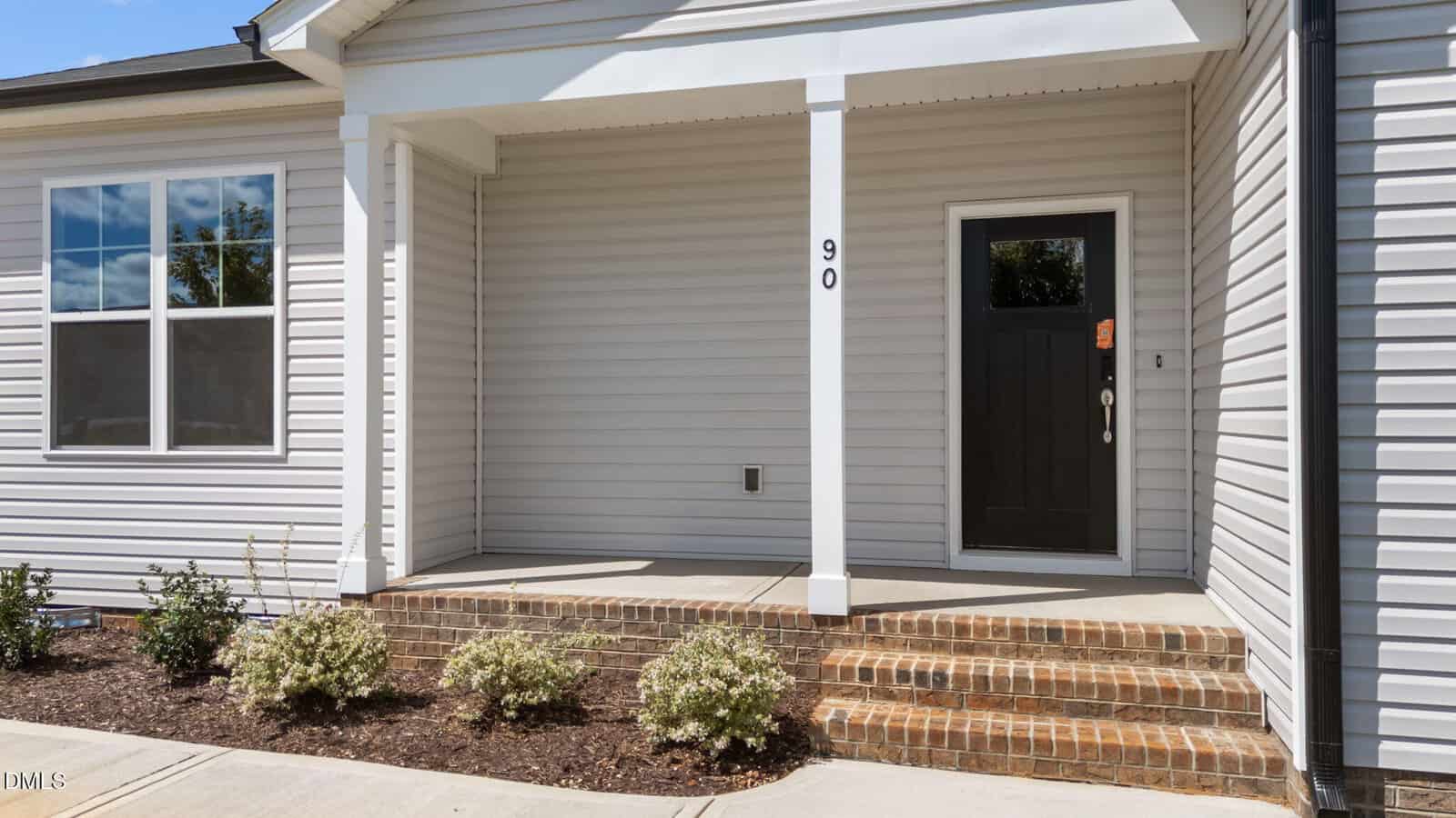 A modern house entryway with light-colored siding, a black door, a covered porch, brick steps, and small shrubs planted along the walkway. The number 90 is displayed next to the door.
