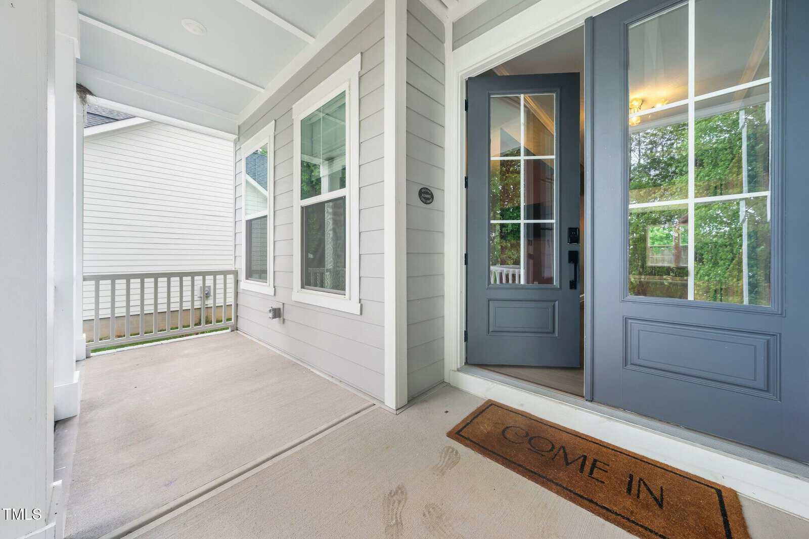 A modern front porch with gray siding, white trim, and a glass-paneled blue door. A welcome mat reading "COME IN" is by the entrance, and greenery is visible through the windows.