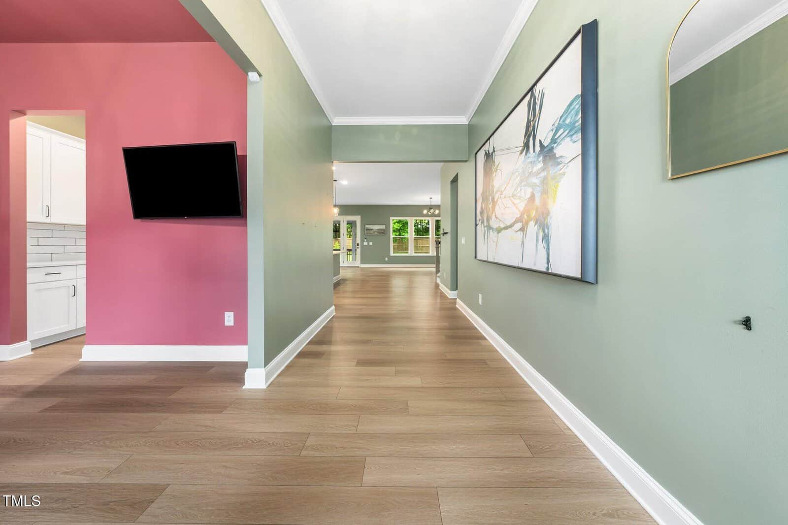 A modern hallway with light wood flooring, green walls, and a large abstract painting. To the left is a kitchen with white cabinets and a pink accent wall. At the end, glass doors open to a bright, green outdoor area.