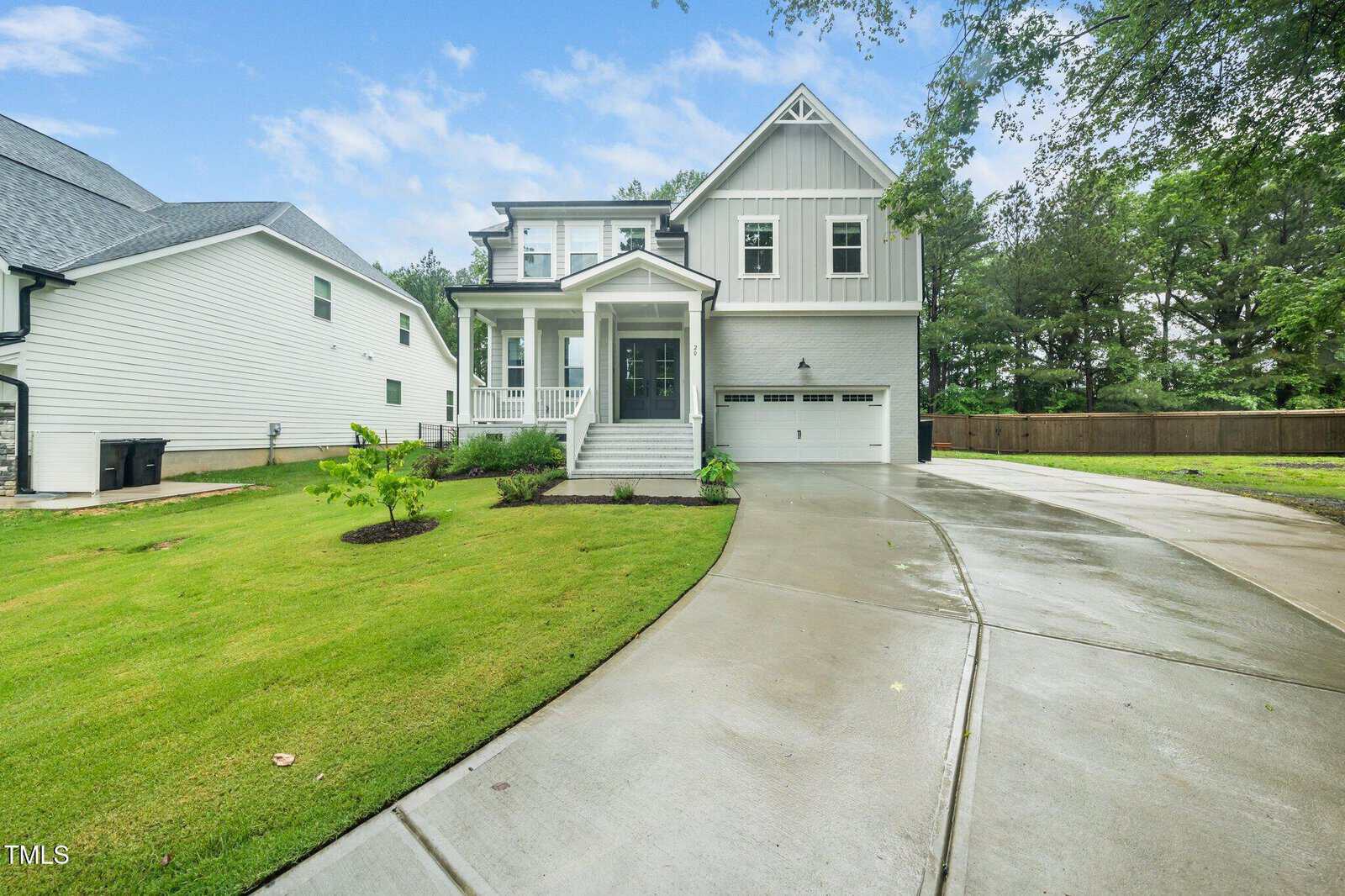 A modern two-story white house with a covered front porch, double garage, and a curved concrete driveway, surrounded by a neat green lawn and trees in the background under a partly cloudy sky.