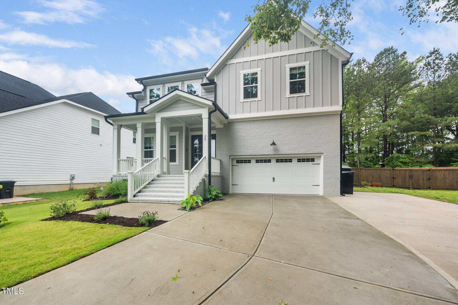 Modern two-story house with light gray siding, white trim, a covered front porch, and a double garage. The home is surrounded by a neat lawn, garden beds, and a concrete driveway on a cloudy day.