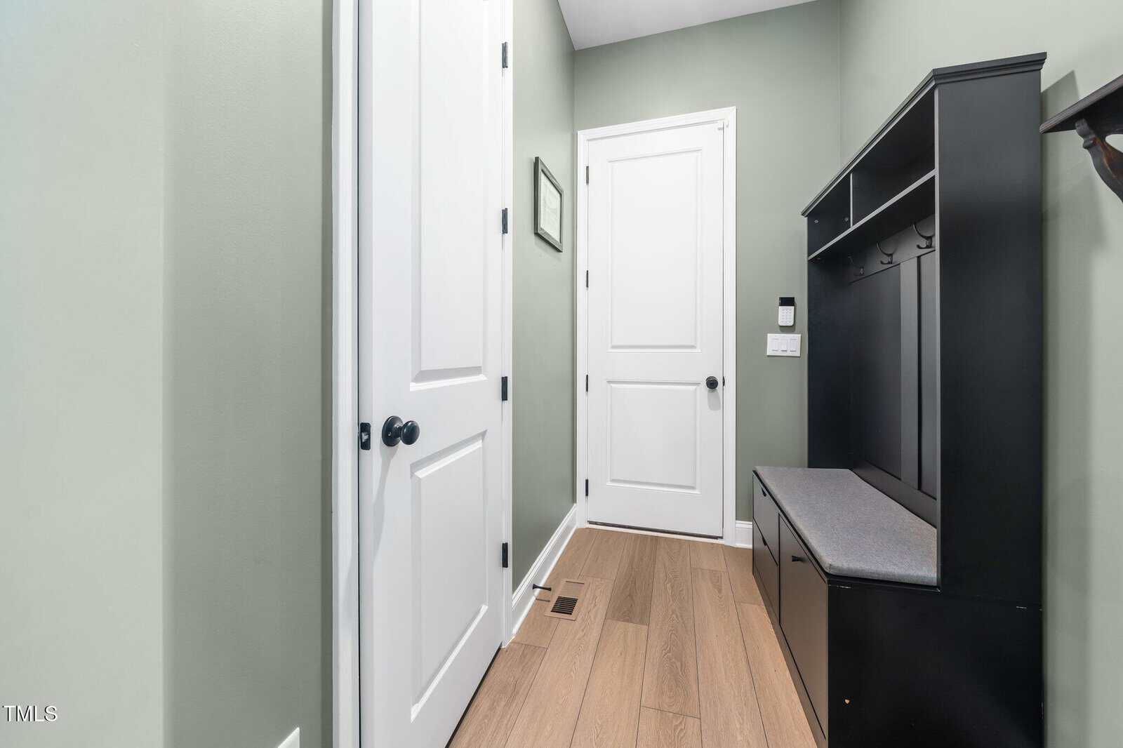 A hallway with light green walls, two closed white doors, wood flooring, and a black hall tree bench with hooks and storage. A vent is on the floor and a small framed picture hangs on the wall.