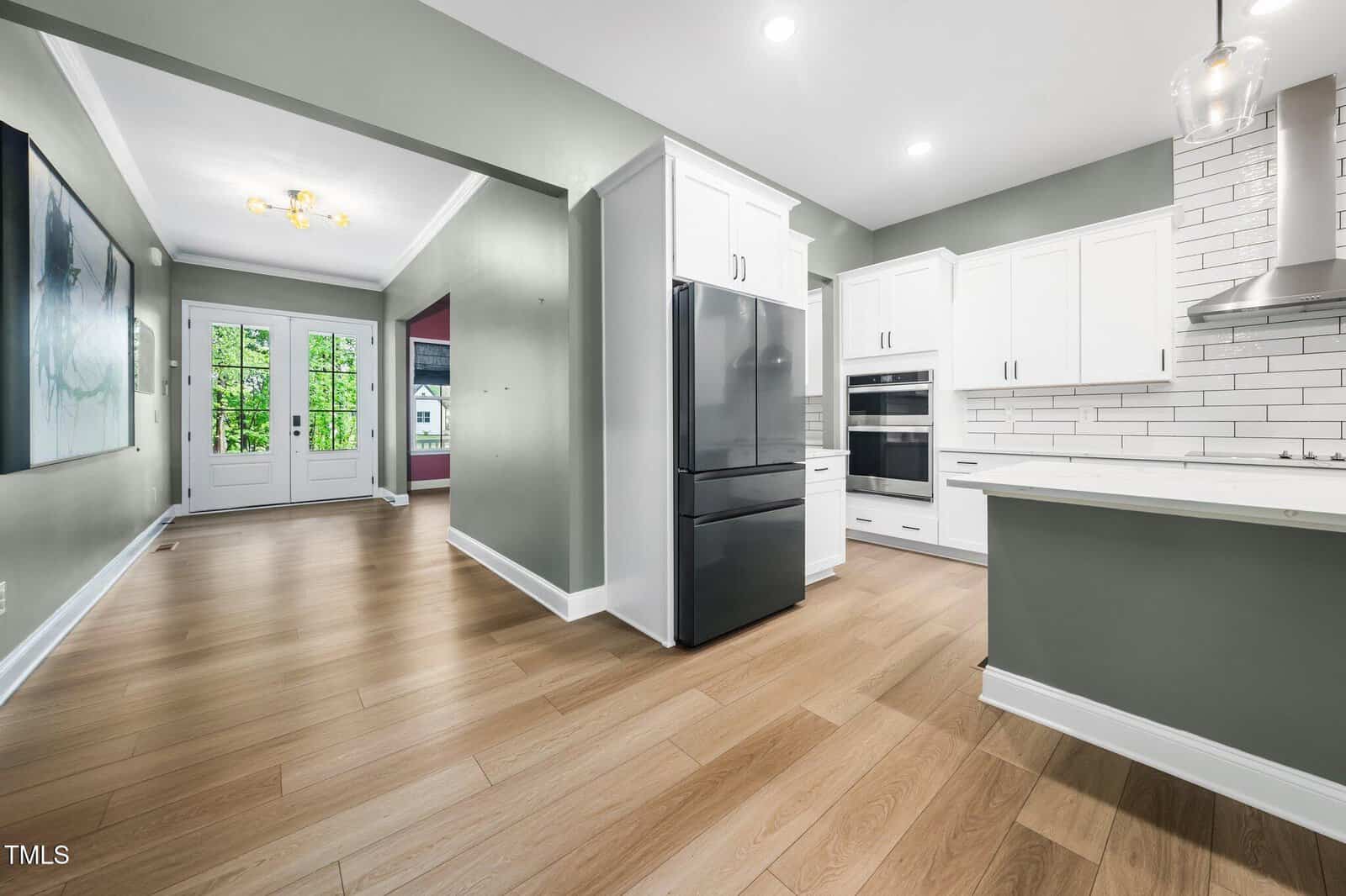 Modern kitchen with white cabinets, black refrigerator, double oven, light wood floors, and gray-green walls; the room opens to a hallway with large windows and double doors leading outside.
