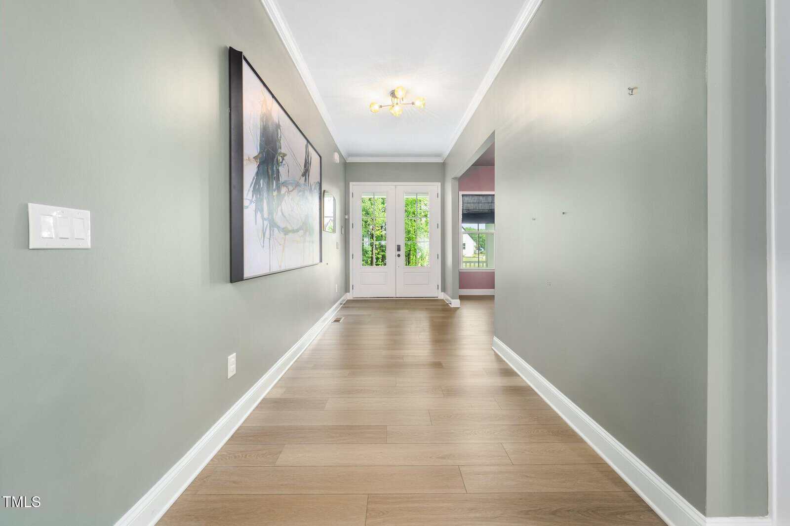A hallway with light wood flooring, pale green walls, crown molding, a large abstract painting on the left wall, and double white doors with glass panels at the far end, letting in natural light.