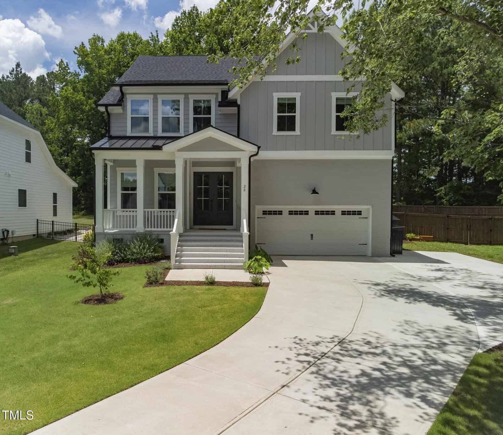 A modern two-story house with light gray siding, a front porch, black roof, and a double garage. The home sits on a green lawn with a curved driveway and trees in the background.