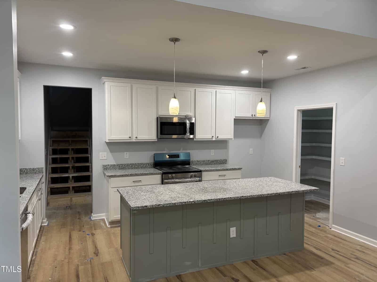 Modern kitchen with white cabinets, stainless steel appliances, granite countertops, an island with pendant lights, wood flooring, and a walk-in pantry to the right. The room is well-lit with recessed ceiling lights.
