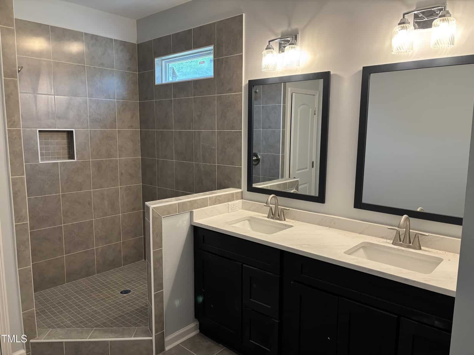 Modern bathroom with a walk-in tiled shower, built-in niche, double vanity with two sinks, white marble countertop, and two wall-mounted mirrors with light fixtures above each. Walls are light-colored with dark cabinetry.