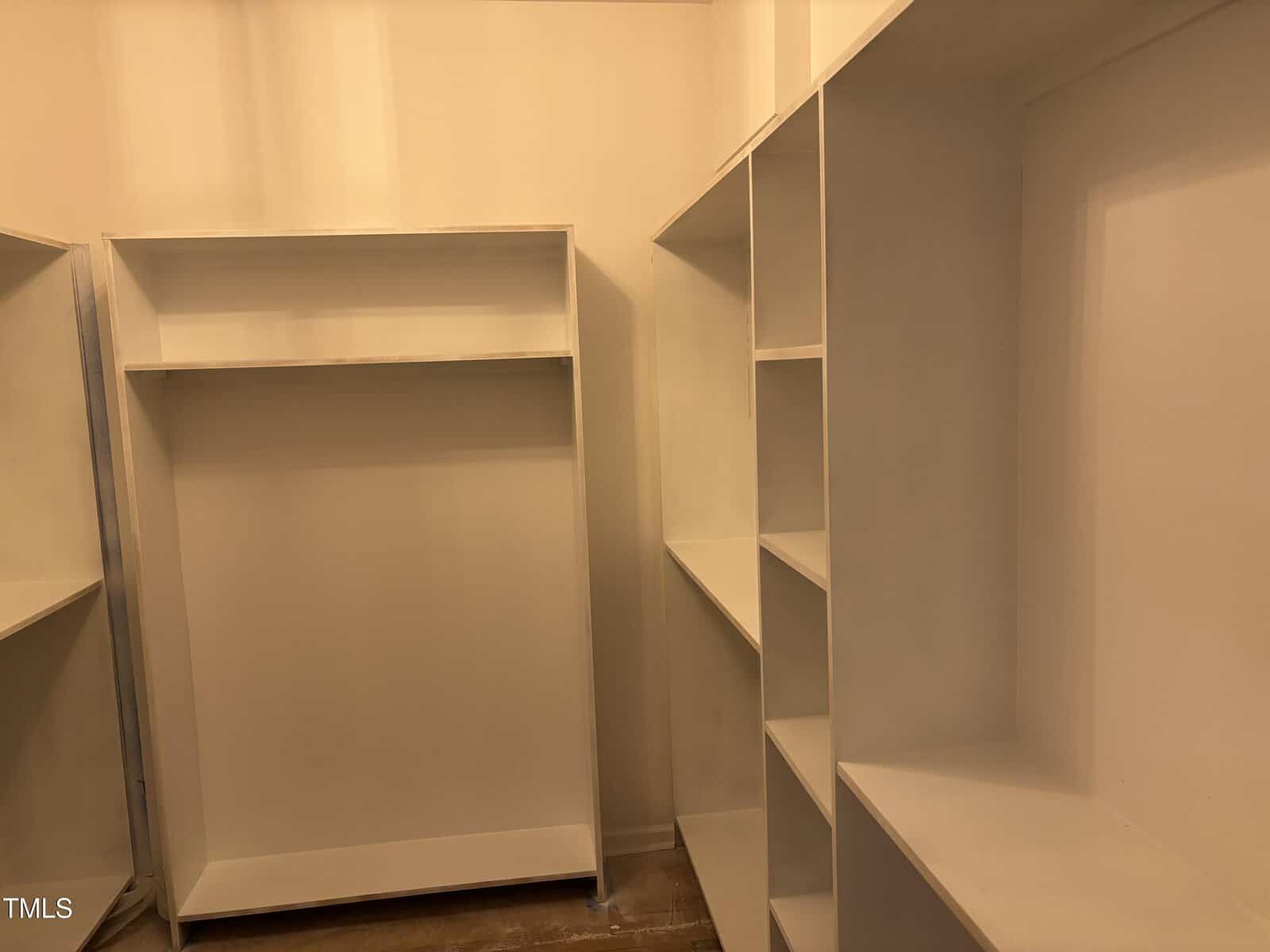 A walk-in closet with empty white shelves and hanging space, featuring built-in storage units on the left and right walls, and a hardwood floor. The lighting casts a warm, yellowish tone.