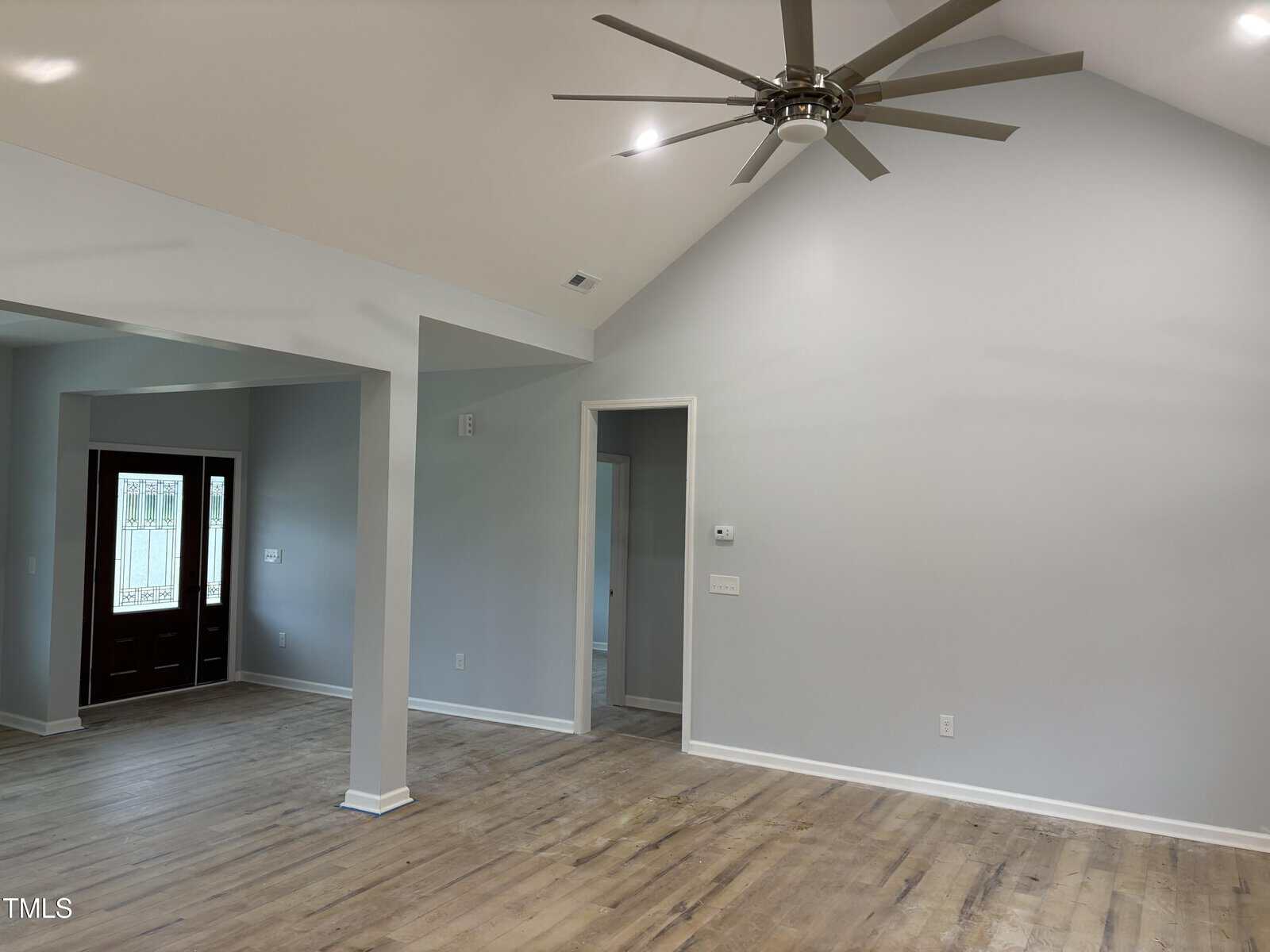 A spacious, empty living room with light wood floors, light gray walls, a vaulted ceiling, and a large ceiling fan. An open doorway leads to the entryway, featuring a dark wood front door with glass panels.
