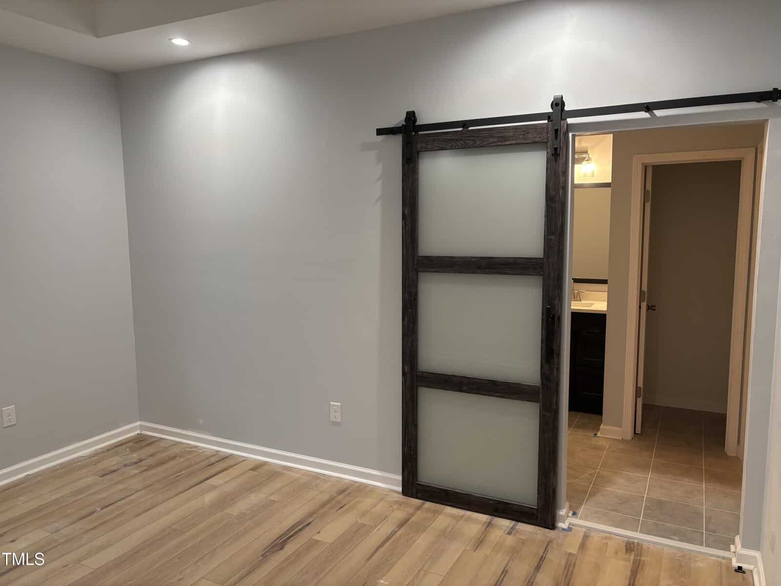 A room with light wood flooring, pale gray walls, and a sliding barn door with frosted glass panels partially open to reveal a tiled bathroom with a vanity and mirror inside.