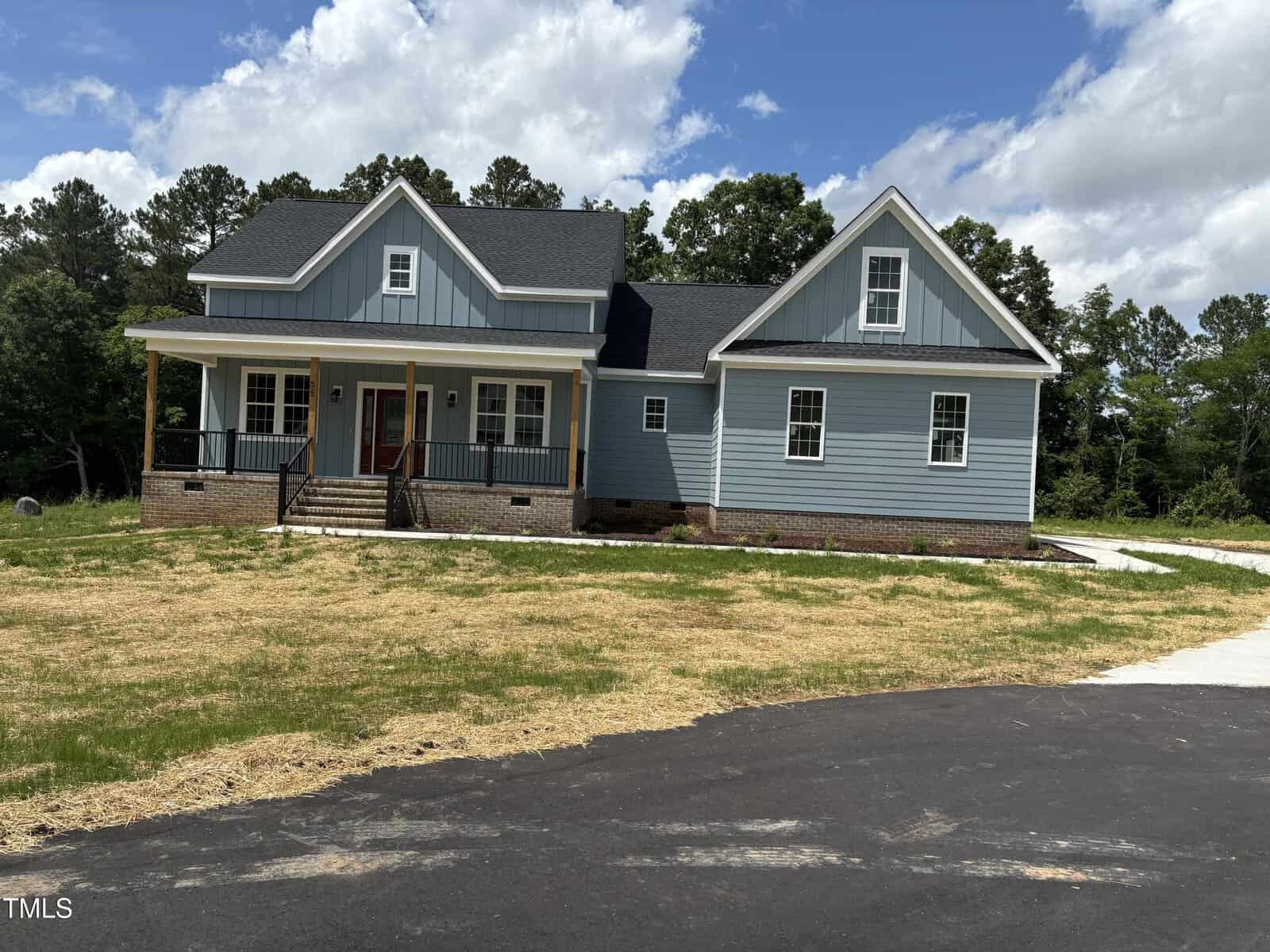 A newly built blue house with white trim, a covered front porch, and multiple gables sits on a yard with fresh straw and a paved driveway, surrounded by trees under a partly cloudy sky.