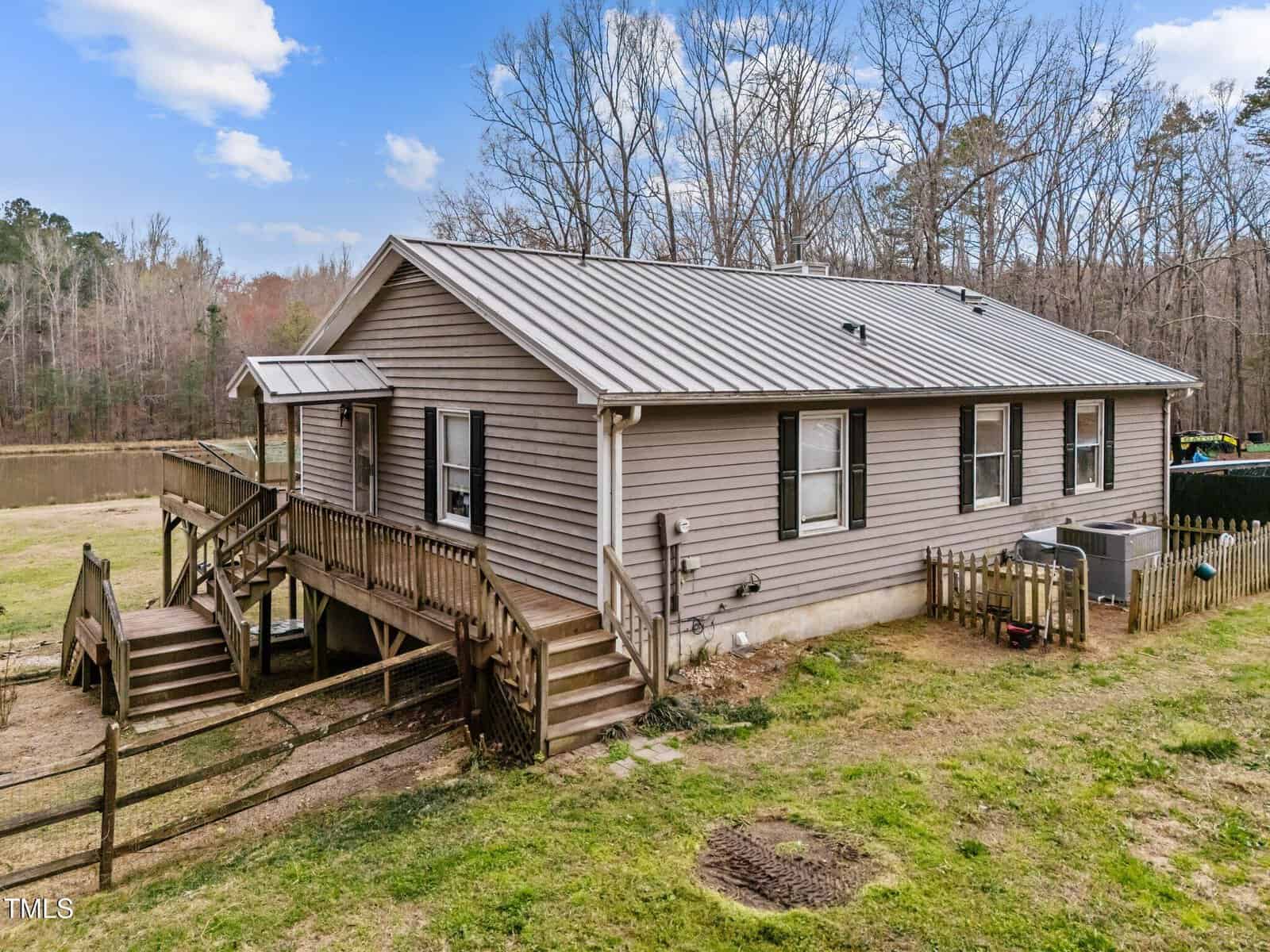 Single-story house with gray siding and a metal roof, elevated on a small foundation with wooden stairs and railing. The yard is partially fenced, and there is a wooded area and pond in the background.