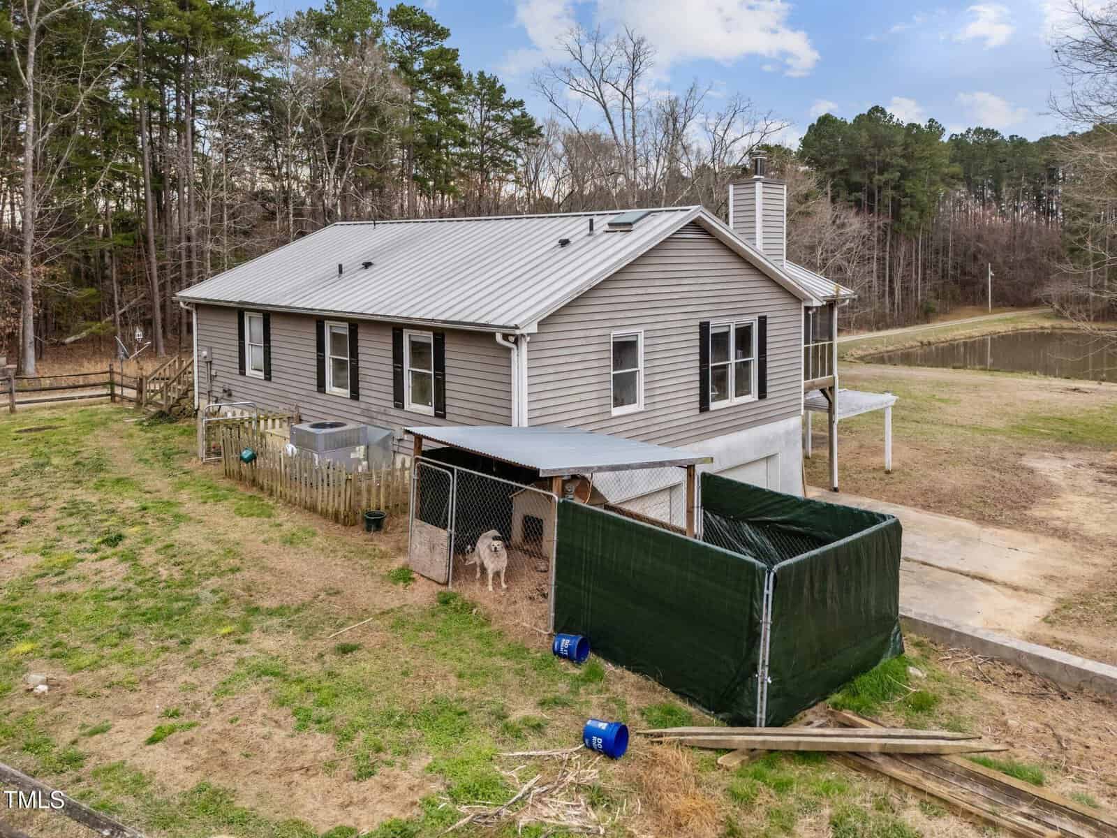 A single-story beige house with a metal roof sits beside a fenced yard area containing a covered dog kennel. Trees surround the property, and a small pond is visible in the background.