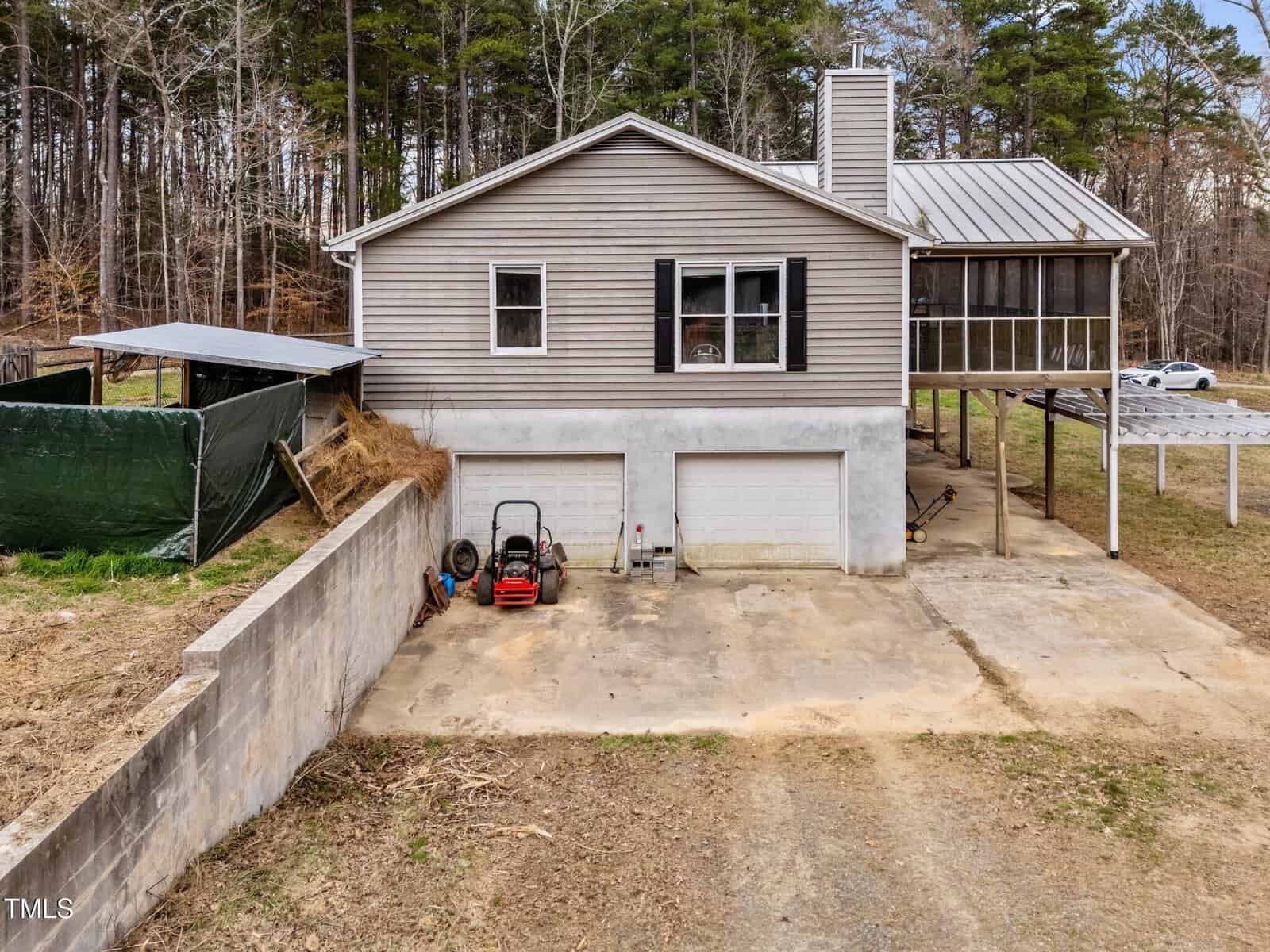A two-story house with gray siding and a metal roof, a double garage on the lower level, a screened porch on the right, and a concrete driveway. A lawn mower, hose reel, and tools are on the driveway. Trees line the background.