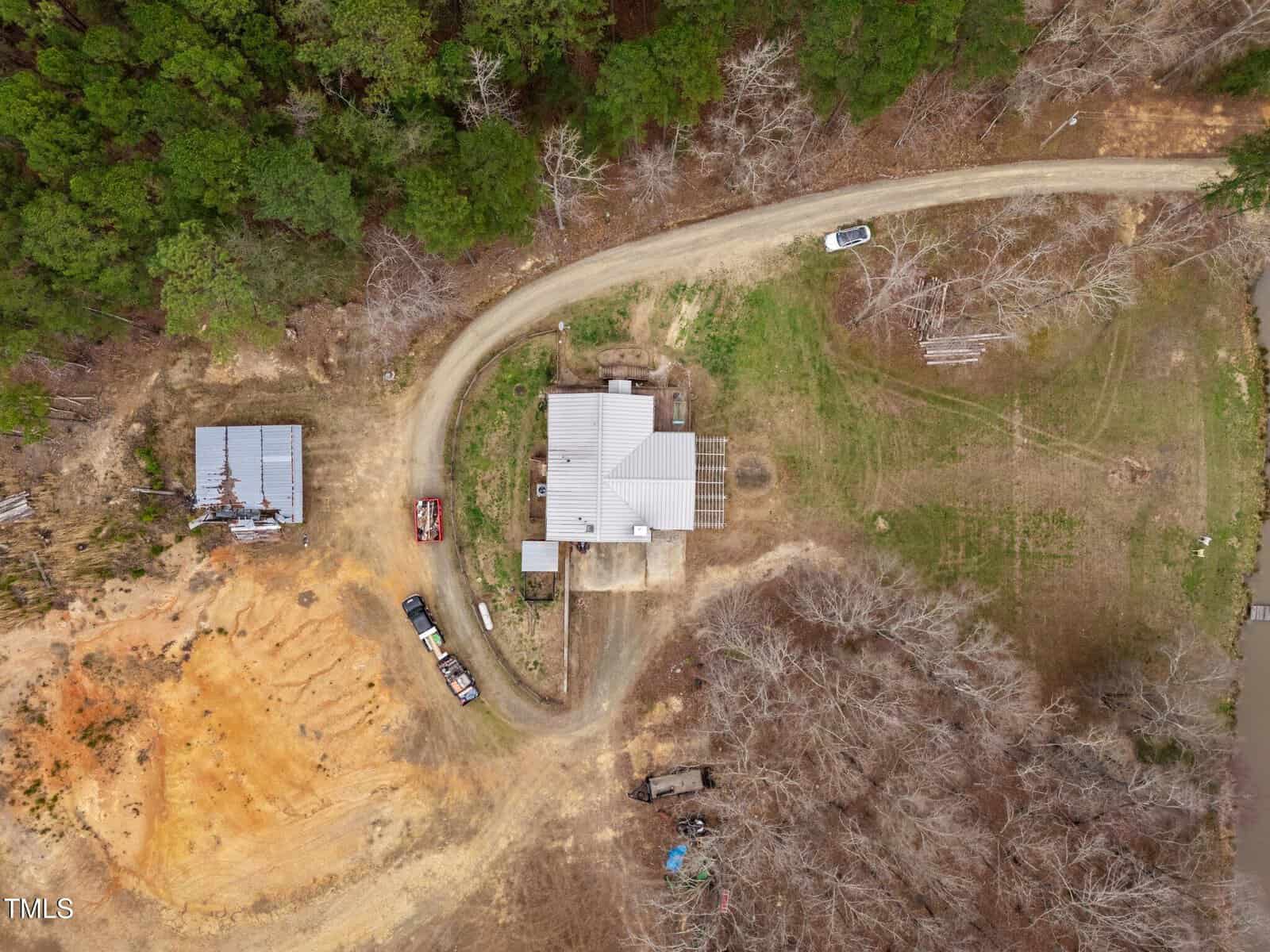Aerial view of a house with a metal roof surrounded by trees, dirt paths, several vehicles, a shed, and a grassy area. The photo shows a mix of cleared and forested land.