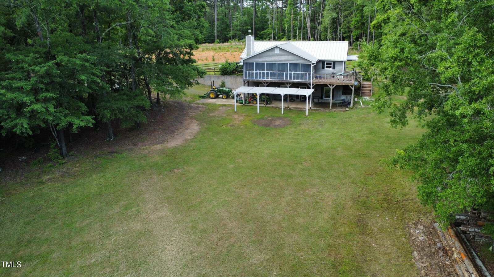 A house with a screened-in porch sits on a large grassy yard, bordered by trees. A covered area with a tractor and other equipment is visible next to the house. Dense forest is seen in the background.