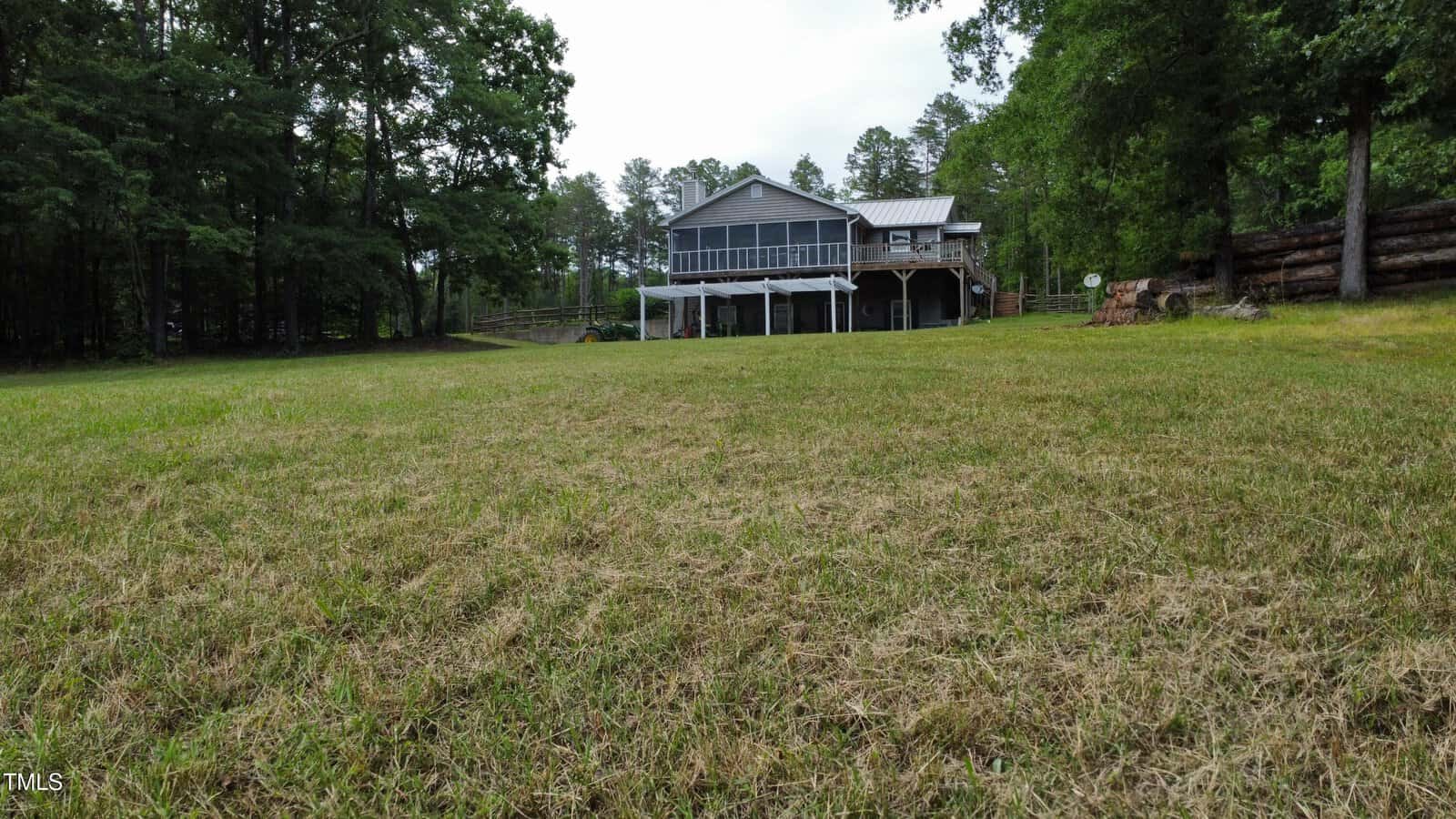 A wide grassy yard leads up to a two-story house with a large screened porch, surrounded by tall trees on both sides. The sky is partly cloudy and the property appears to be in a rural setting.