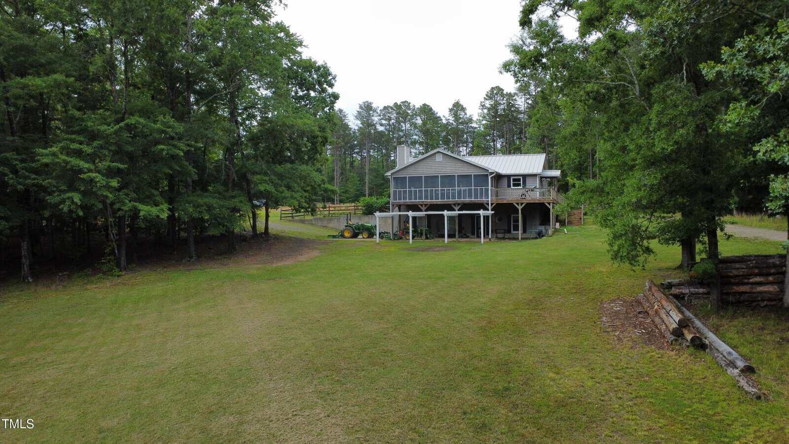 A two-story house with a large screened porch sits on a spacious grassy lawn, surrounded by tall trees. There is a tractor parked beside the house and a stack of logs on the right side of the image.
