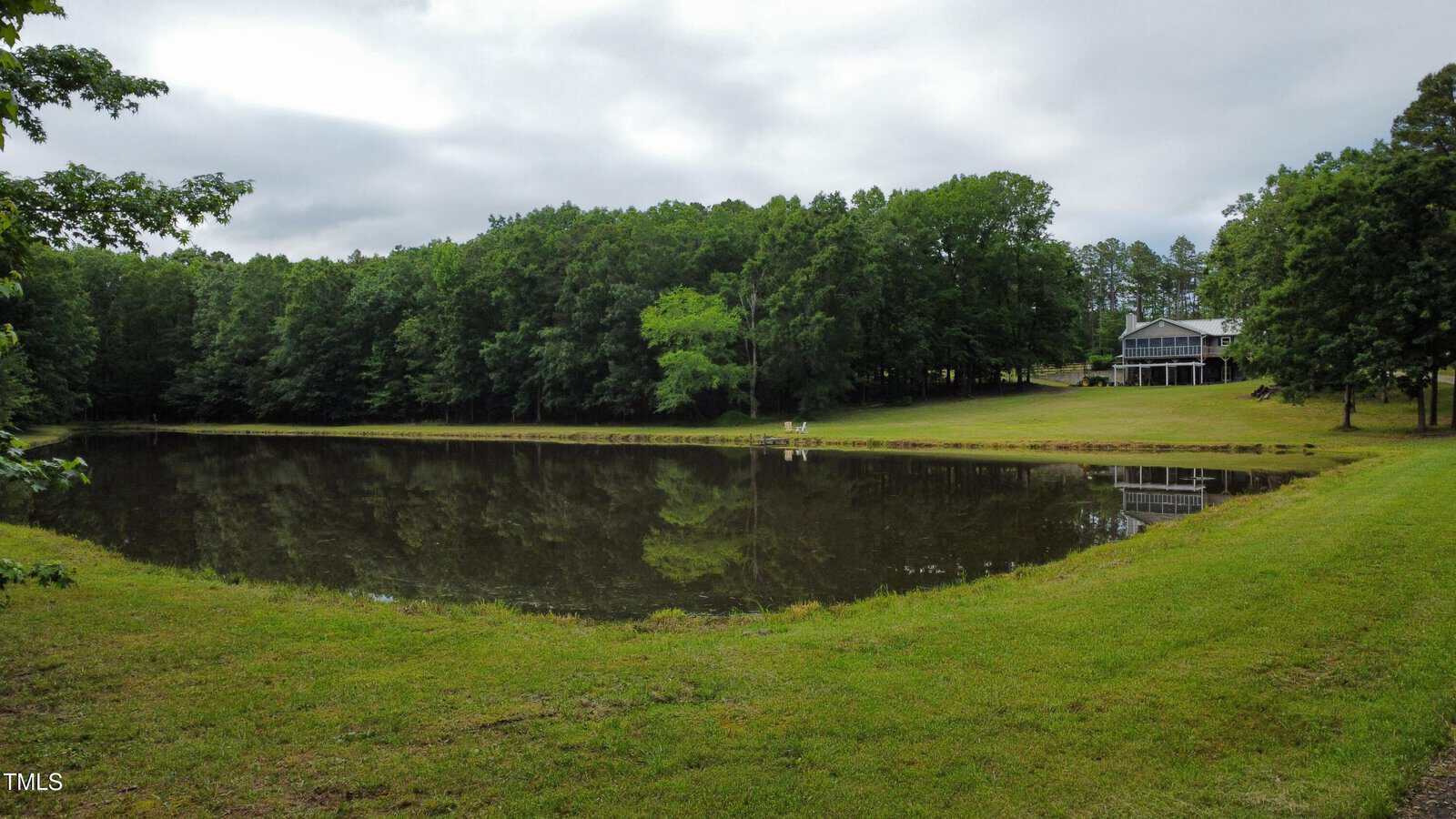 A small pond surrounded by green grass and dense trees with a house and porch visible in the background under a cloudy sky.