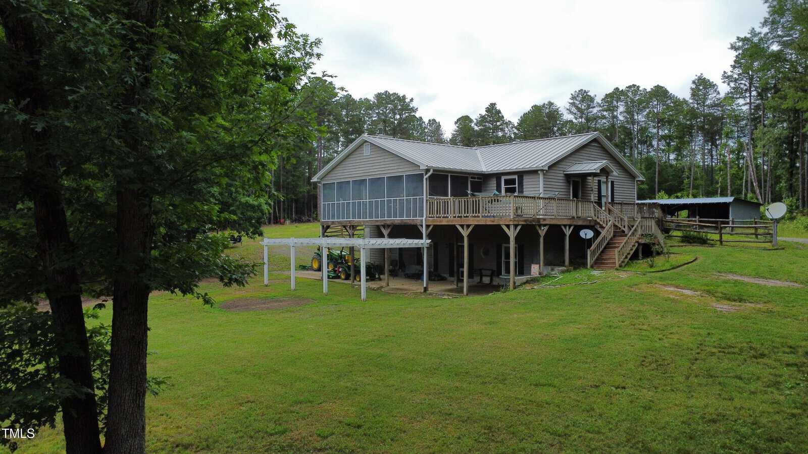 A raised house with a metal roof, screened porch, and wooden deck sits on a grassy lot surrounded by trees. Stairs lead to the main entrance, and there is covered parking space beneath the house.