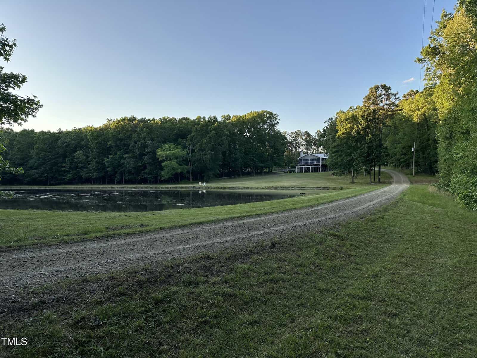 A gravel road curves around a small pond bordered by grass and trees, leading up to a white house with a covered porch at 501 Enon Road, Oxford, set among dense woods under a clear blue sky.