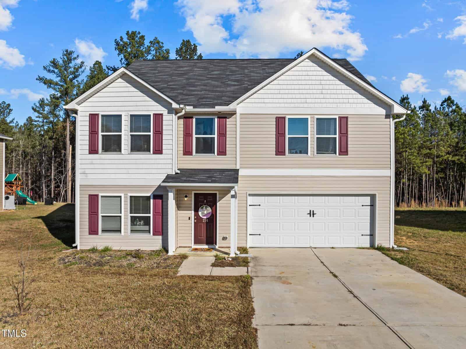 Two-story beige house with white trim, red shutters, and a gray shingle roof. Attached two-car garage with a white door. Concrete driveway and a patchy front lawn. Trees line the background under a partly cloudy sky.
