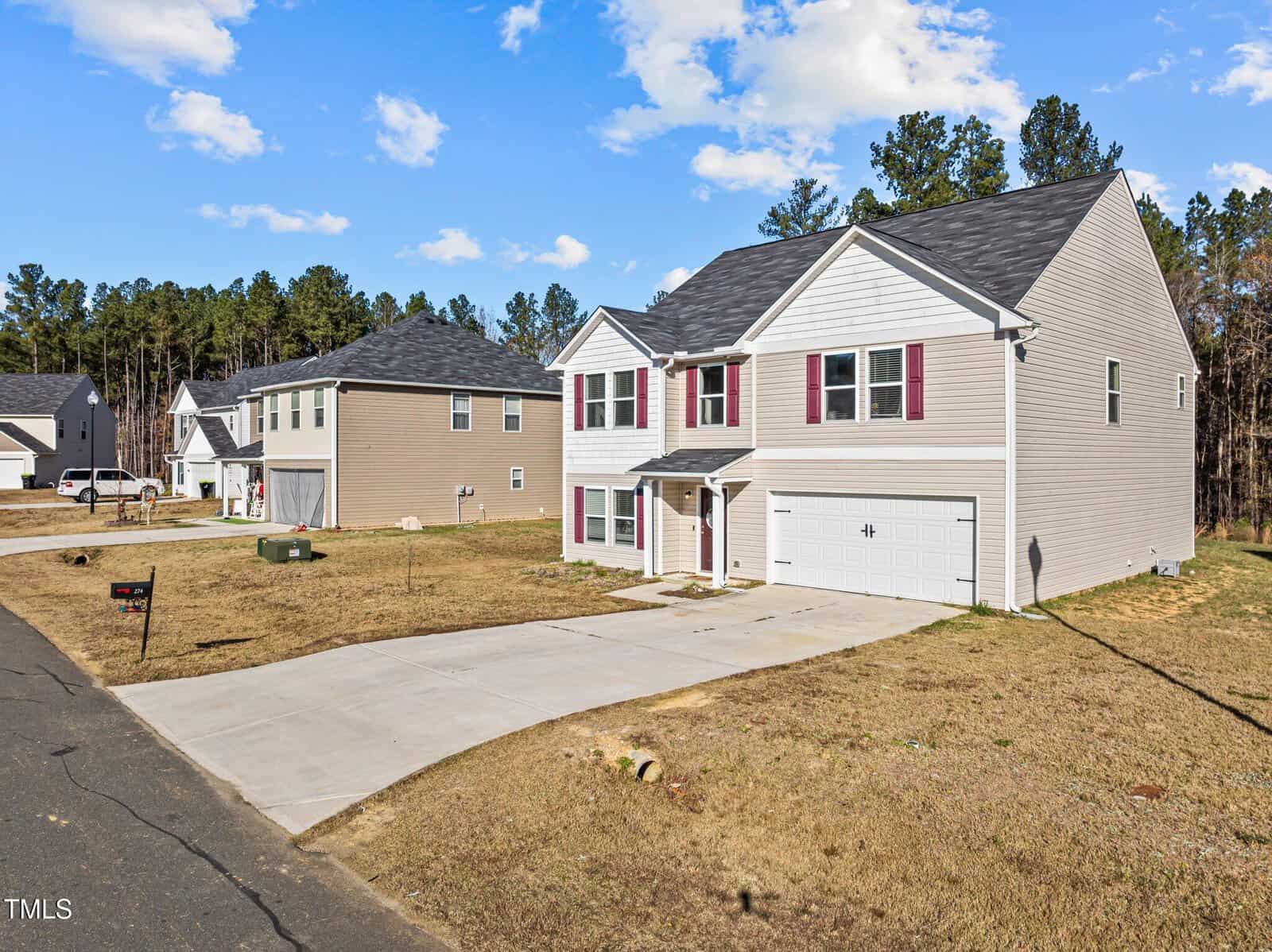 A two-story suburban house with gray siding, red shutters, and a two-car garage, situated on a spacious lot with a large driveway and minimal landscaping in a residential neighborhood.