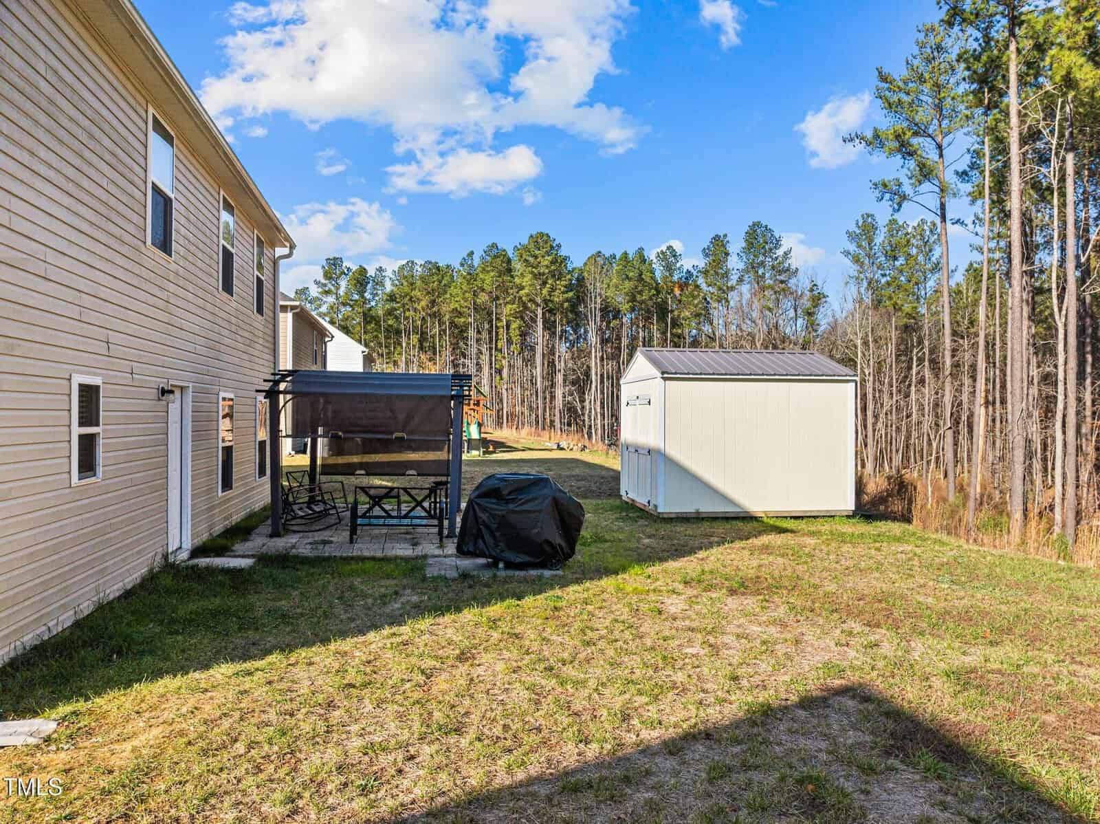 A backyard with a tan two-story house on the left, a covered patio set and grill in the center, a white storage shed in the background, and a wooded area beyond the grassy yard.