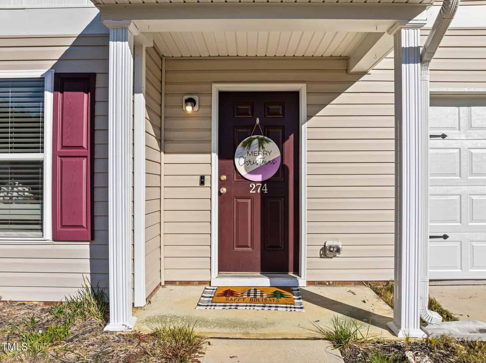 A maroon front door with a “Merry Christmas” sign, a “Happy Holidays” doormat, and white columns on a beige house. The door has the number 274 above the handle. Grass and concrete are in front.