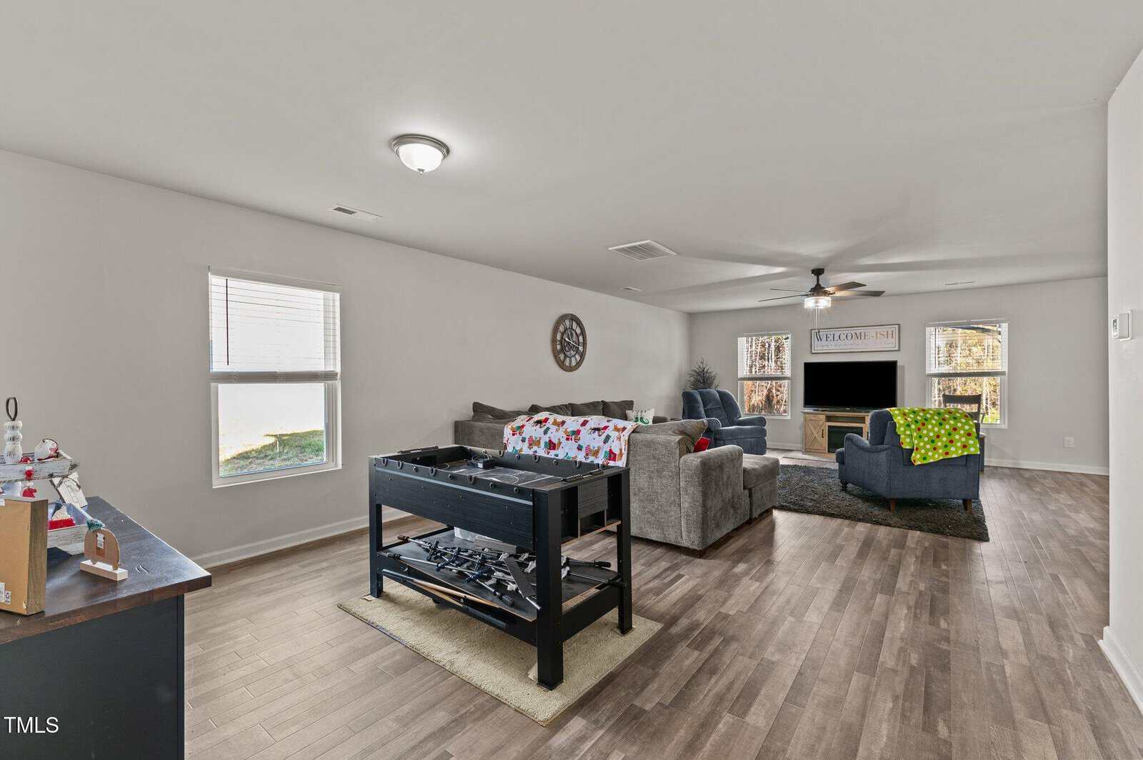 Spacious living room with wood flooring, gray sofas, a foosball table, large TV, and holiday decor. Two windows let in natural light. A ceiling fan and wall clock are visible.