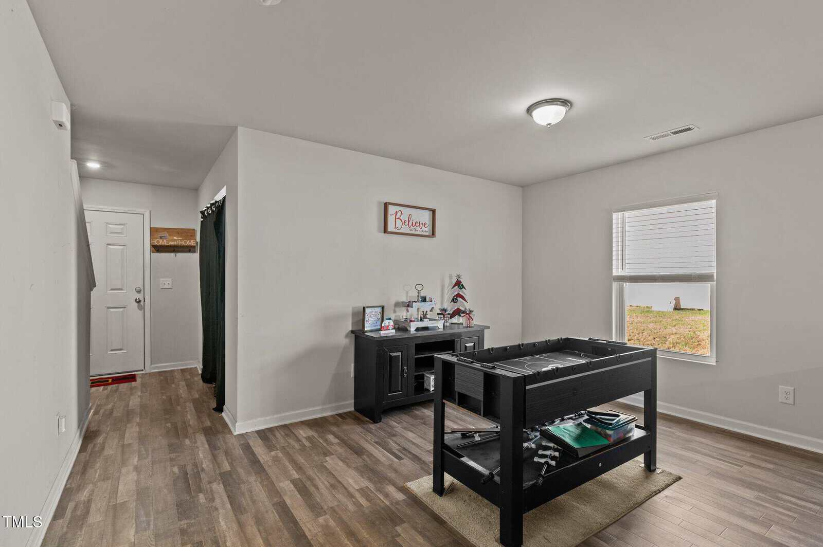 A room with wood flooring, a foosball table, a desk with decor, a window, and a small entryway with a door. A “Believe” sign hangs on the wall above the desk.