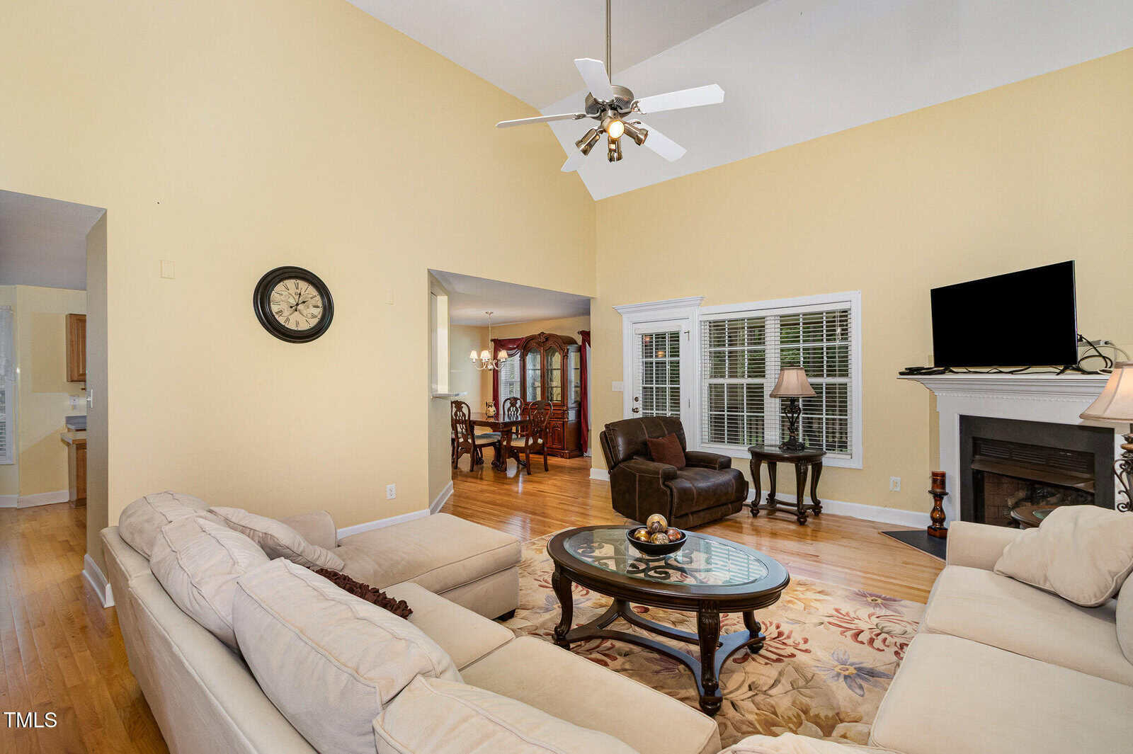 Living room with beige walls, white trim, sectional sofa, coffee table, armchair, area rug, fireplace with TV, ceiling fan, clock on wall, and open view into dining area with table and chairs. Hardwood floors throughout.
