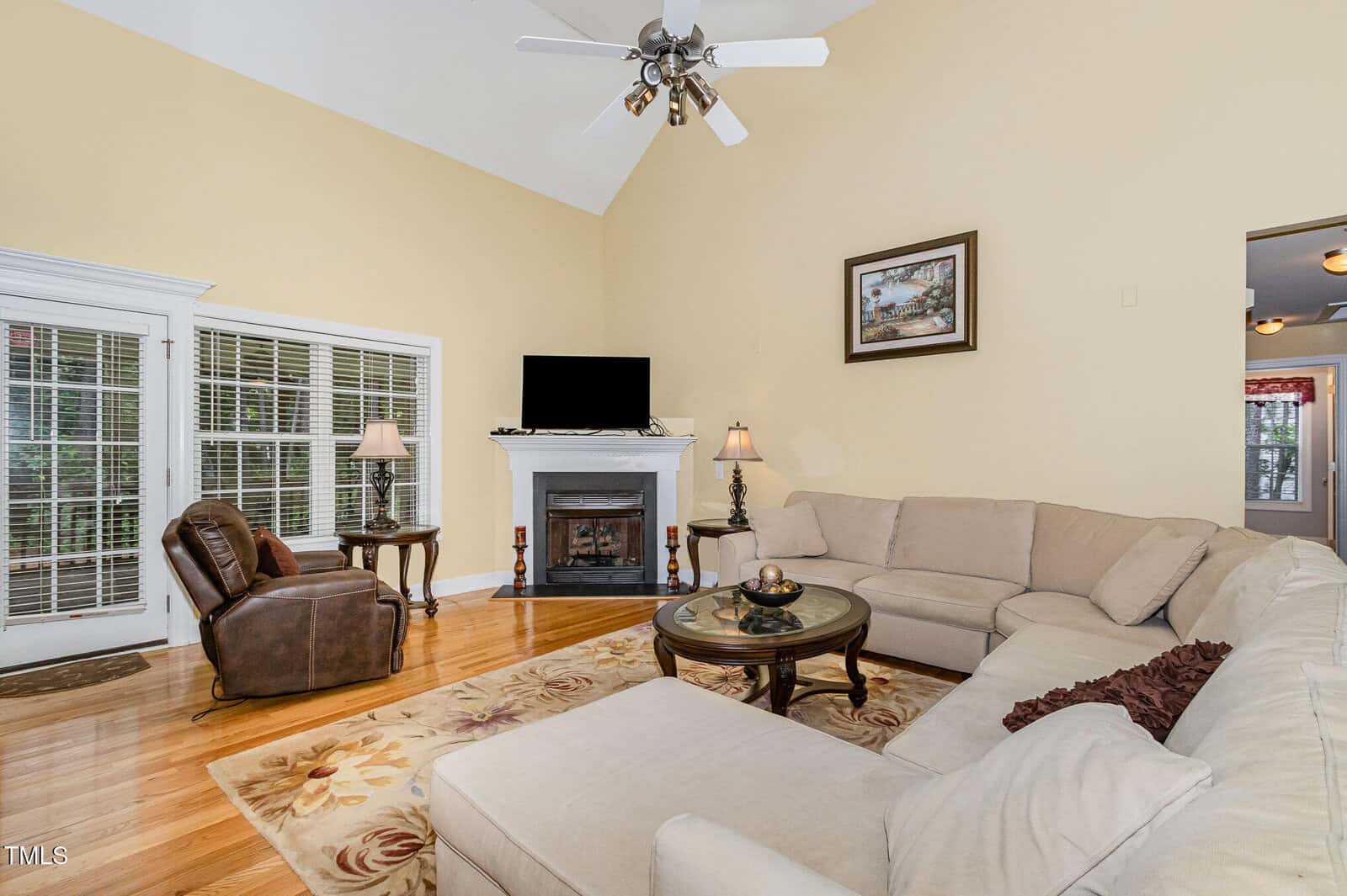A living room with a beige sectional, round coffee table, brown recliner, fireplace with a TV, framed art above mantel, lamps, hardwood floors, French doors, and a ceiling fan under a vaulted ceiling.