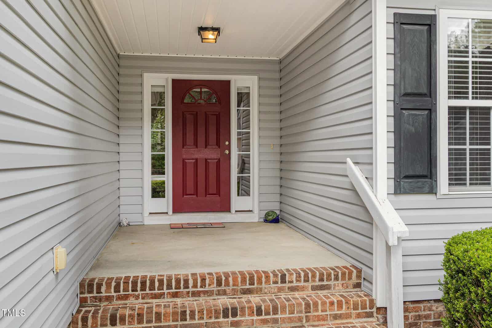 Covered front porch with a red door, sidelights, and brick steps. Gray vinyl siding, white trim, black shutters, and a small shrub are visible. Ceiling light is installed above the entrance.