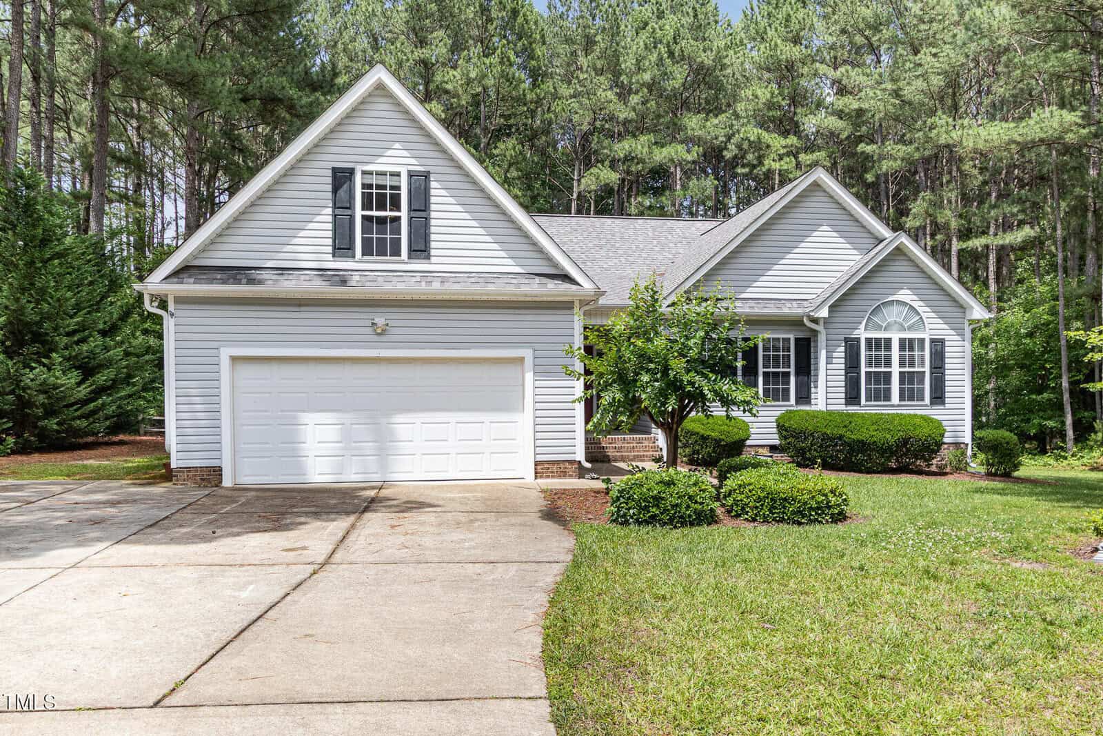 A light gray single-story house with white trim, double garage, arched window, and neatly trimmed bushes in front, surrounded by tall pine trees and a well-maintained lawn.