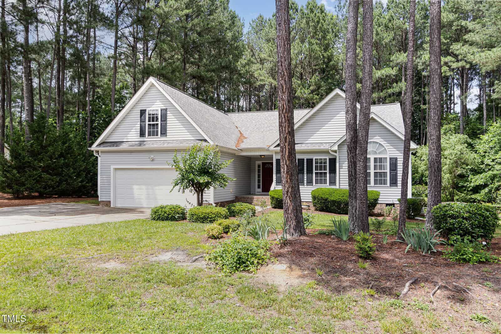 A light gray suburban house with a two-car garage, front porch, and manicured lawn, surrounded by tall pine trees and various shrubs on a sunny day.