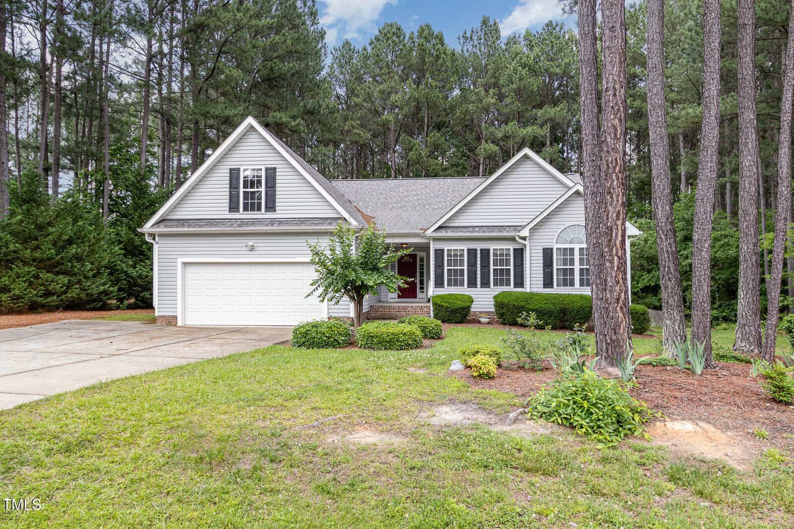 A gray, single-story house with a gabled roof and attached two-car garage sits among tall pine trees, with green shrubs and a manicured lawn in front. A concrete driveway leads up to the garage.