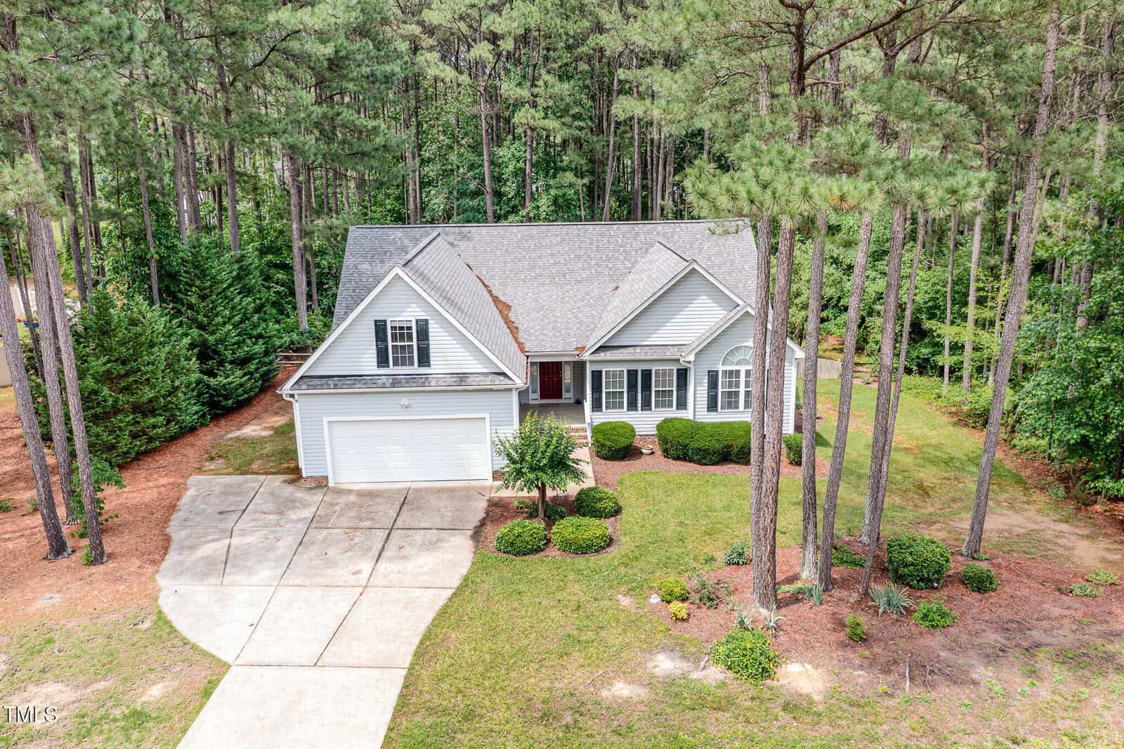 A light gray, single-story house with a two-car garage sits among tall pine trees, surrounded by well-kept grass and shrubs. A curved driveway leads to the front entrance, and a wooded area is visible in the background.