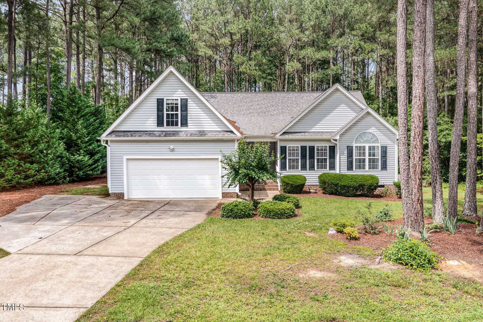 A single-story gray house with white trim, an attached two-car garage, and front shrubs, set against a backdrop of tall pine trees and surrounded by a grassy lawn and concrete driveway.
