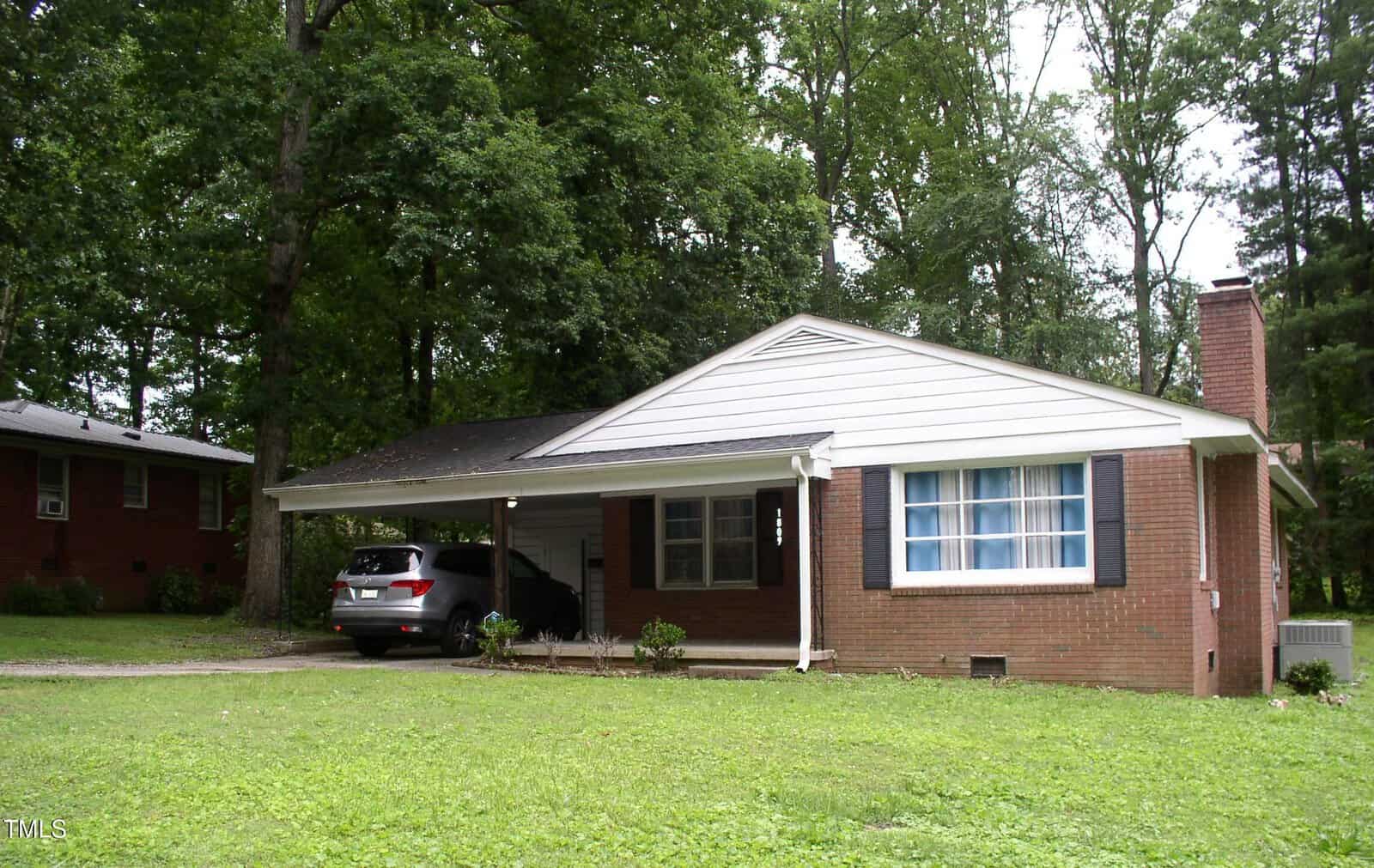 A single-story brick house with white siding at 1809 S Garnett Extension in Henderson features a front porch, blue shutters, a car parked in the covered carport, a grassy yard surrounded by trees, and a chimney on the right side.