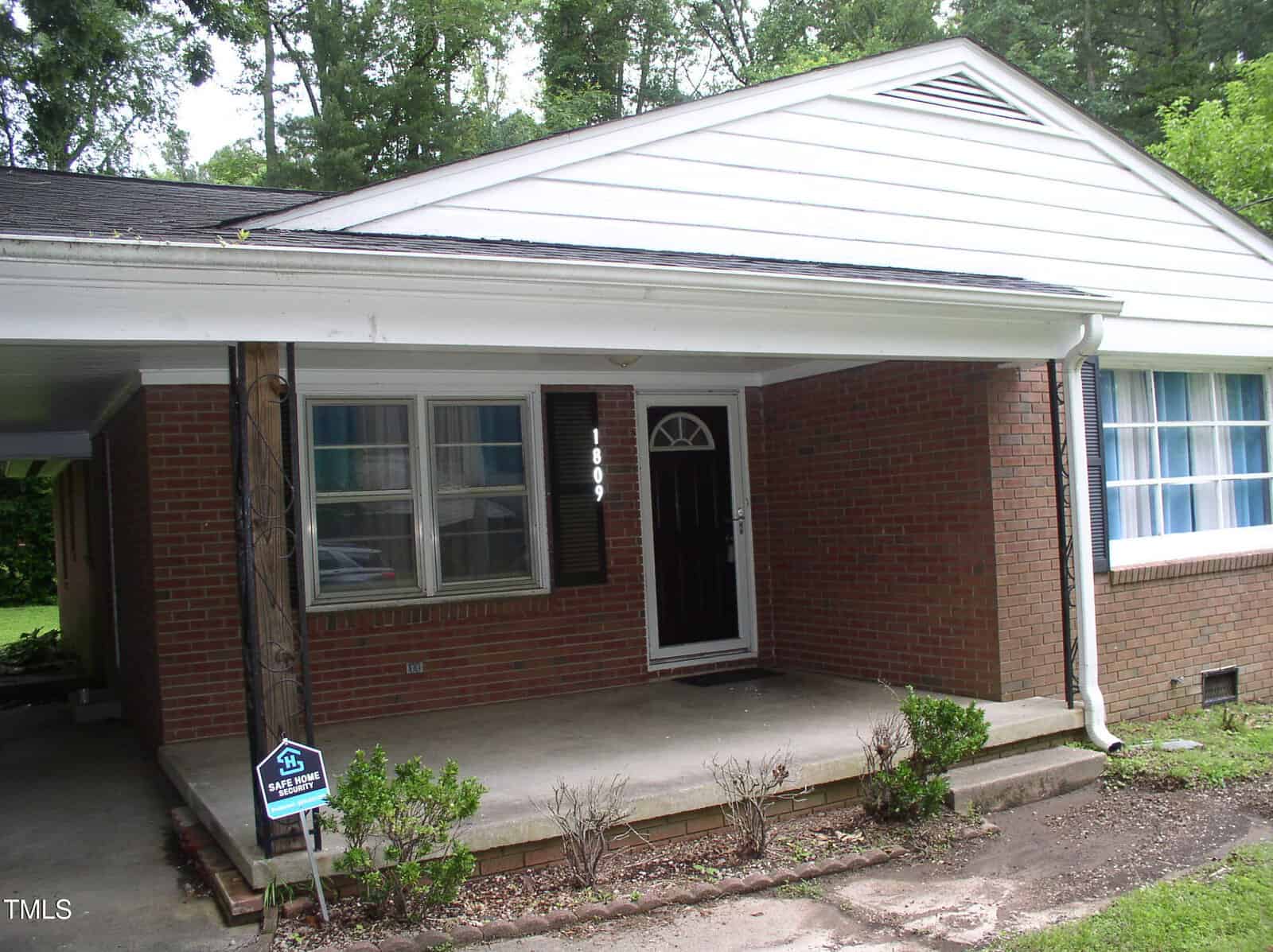 A brick house with white trim features a covered front porch, a black front door, two windows with shutters, and a small yard with minimal landscaping. A carport is attached to the left side.