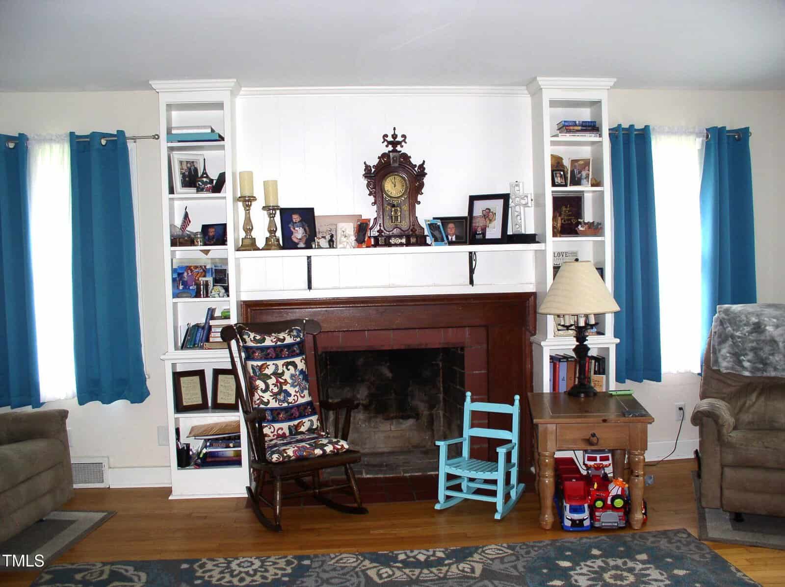A living room with a brick fireplace, shelves with books and photos, blue curtains, a floral rocking chair, a small blue chair, a lamp on a side table, and children’s toys on the wooden floor.