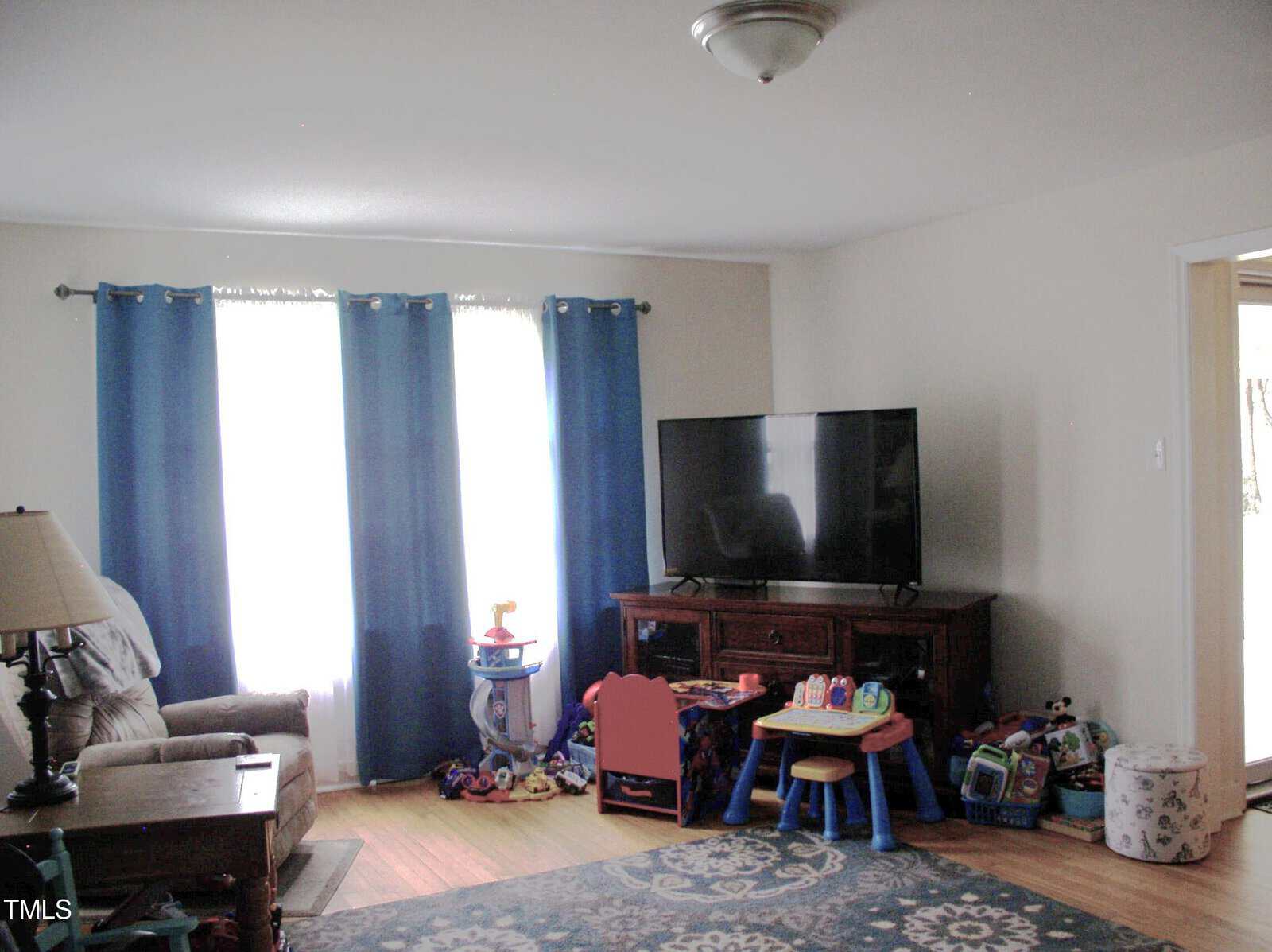 A living room with light walls, blue curtains, a large TV on a wooden stand, children's toys and furniture, an armchair, a side table with a lamp, and a patterned rug on a hardwood floor.