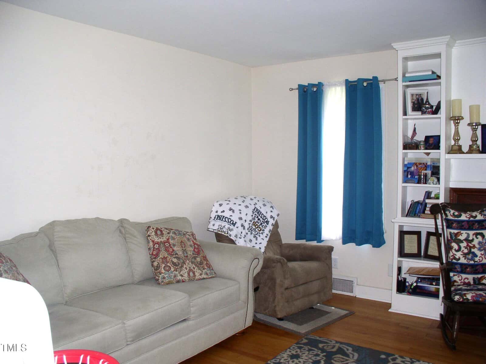A living room with a beige sofa, patterned pillows, a brown recliner with a blanket, blue curtains on a window, hardwood floor, and built-in shelves holding books, photos, and decorative items.