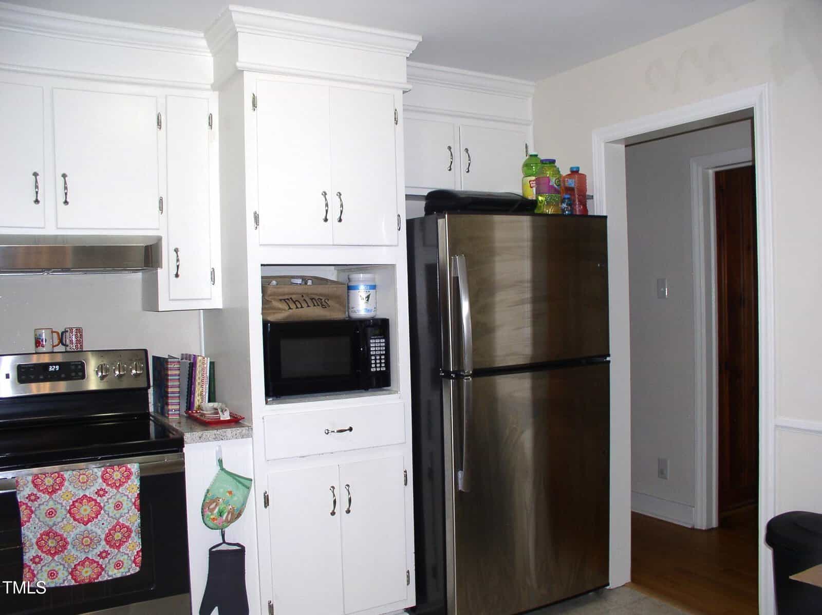 A kitchen with white cabinets, a stainless steel refrigerator, a microwave in a built-in shelf, a black stove with a colorful towel, and various items on top of the fridge and counter.