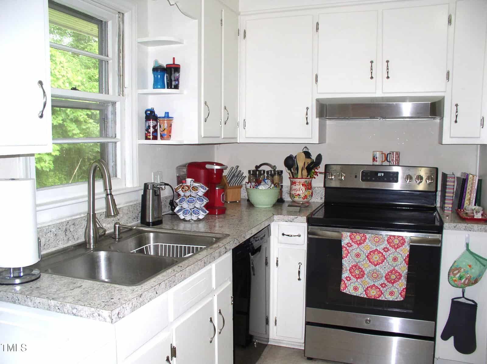 A kitchen with white cabinets, a marble-patterned countertop, an electric stove with a floral towel, a double sink, various utensils, small appliances, and a window with a view of green trees.