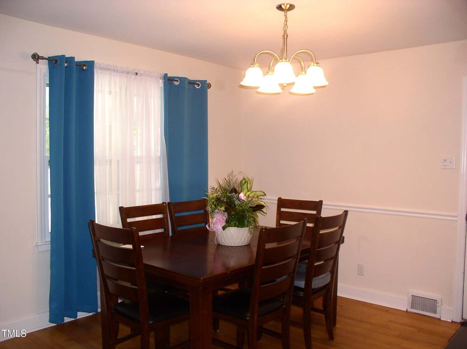 A dining room with a dark wood table and six matching chairs, a potted plant centerpiece, blue curtains on a window, light-colored walls, and a ceiling light fixture with five bulbs.