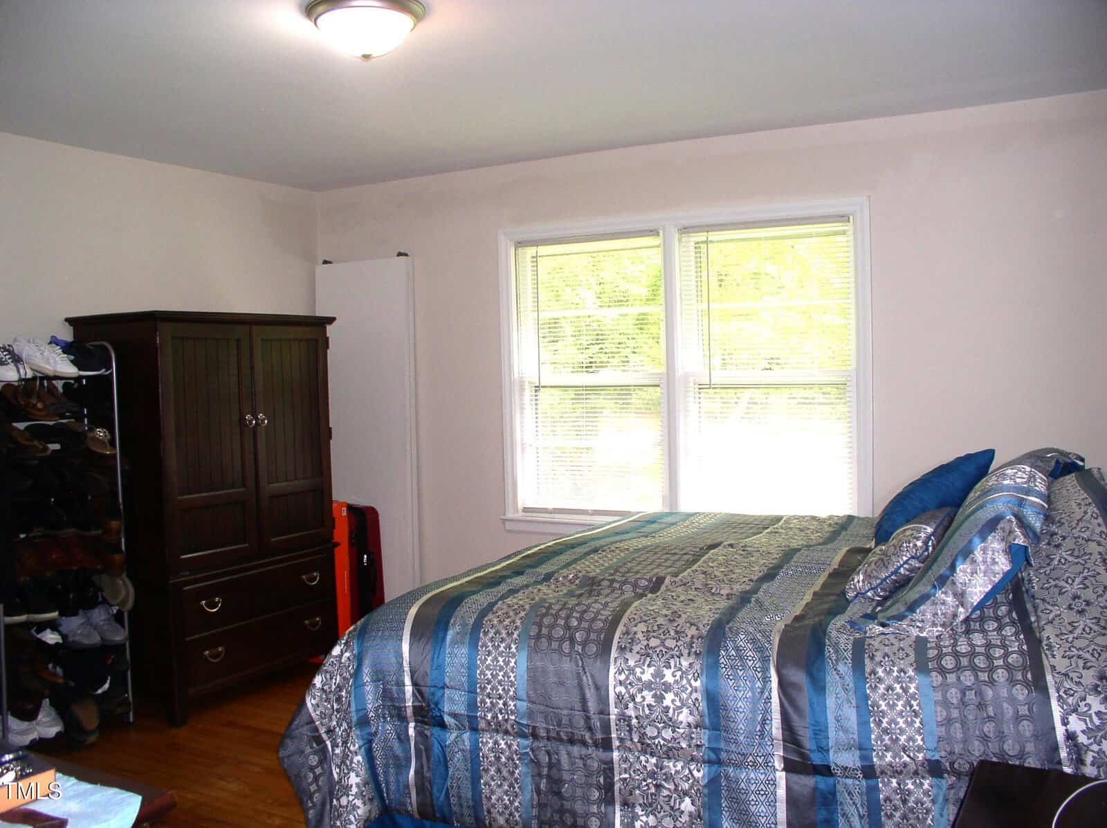 Bedroom with a double bed in blue patterned bedding, a dark wood armoire, shoe rack, hardwood floors, and a window letting in natural light. Walls are light-colored, and the room appears tidy and well-lit.