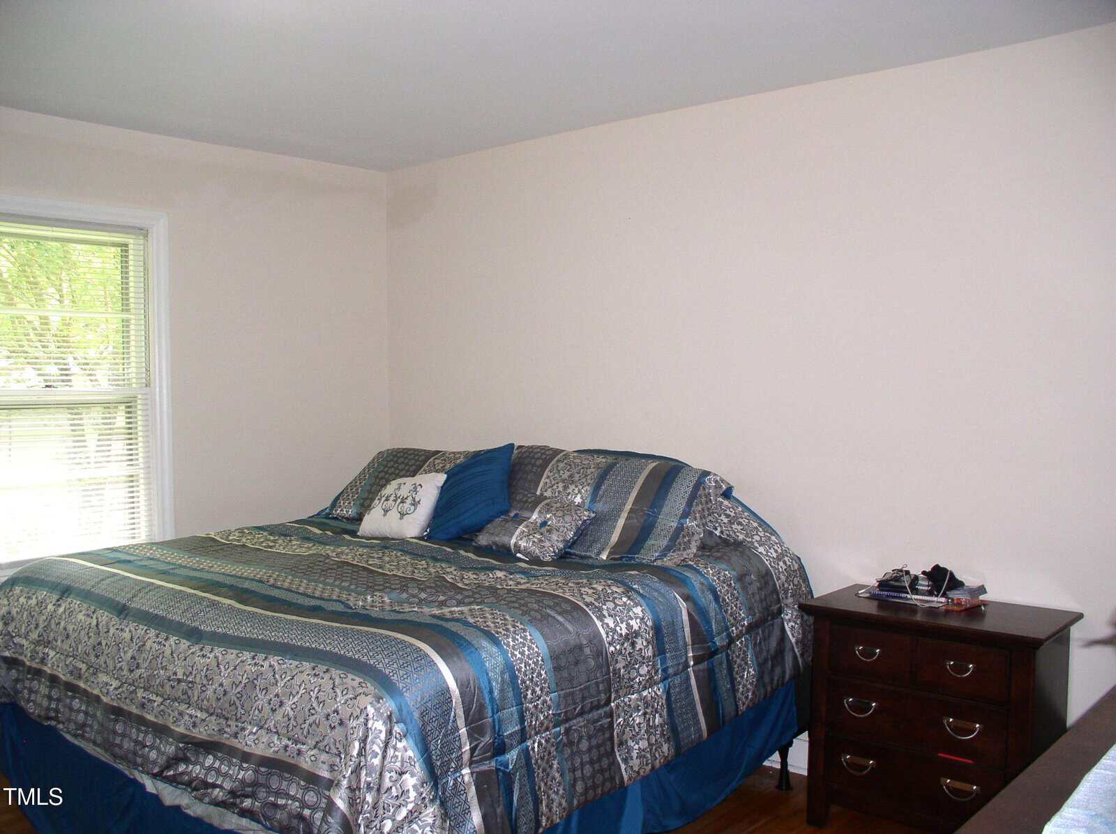 A bedroom with a neatly made bed in blue and gray bedding, a window with blinds, a small wooden nightstand with drawers, and some items on top. Walls are light-colored and the floor is wooden.